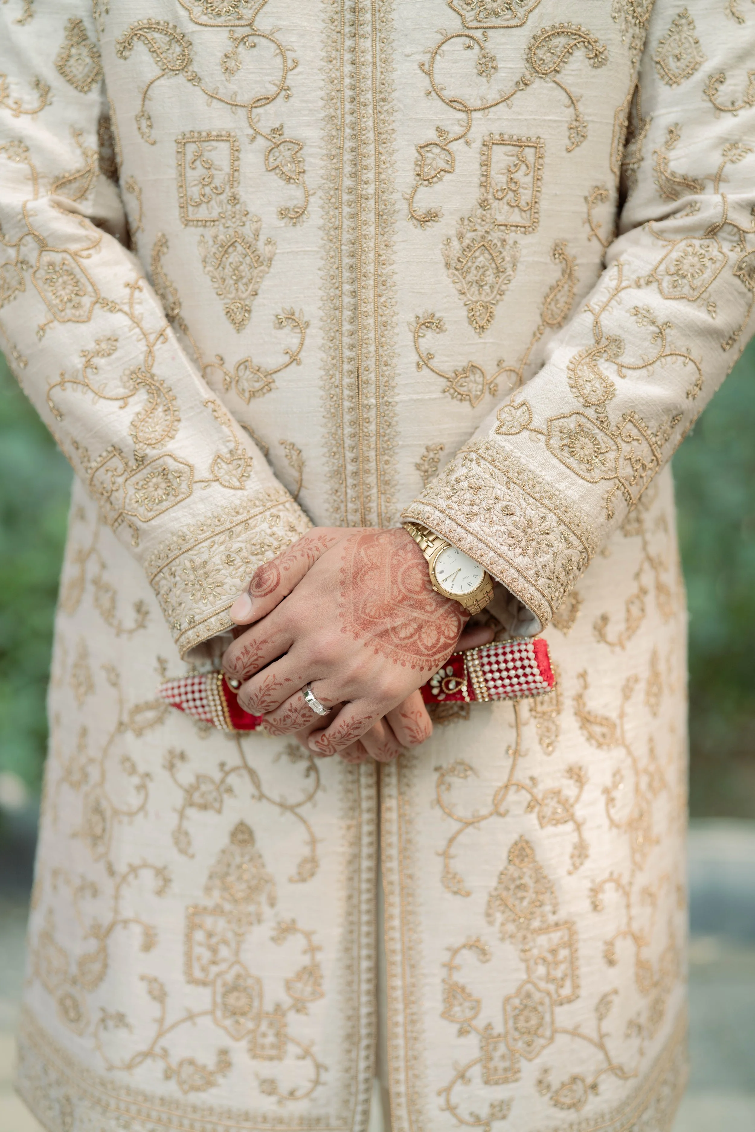 Close-up of a person wearing a cream-colored embroidered traditional outfit with gold thread, holding hands in front of their body, wearing a gold wristwatch and a diamond ring, with henna designs on their hand.