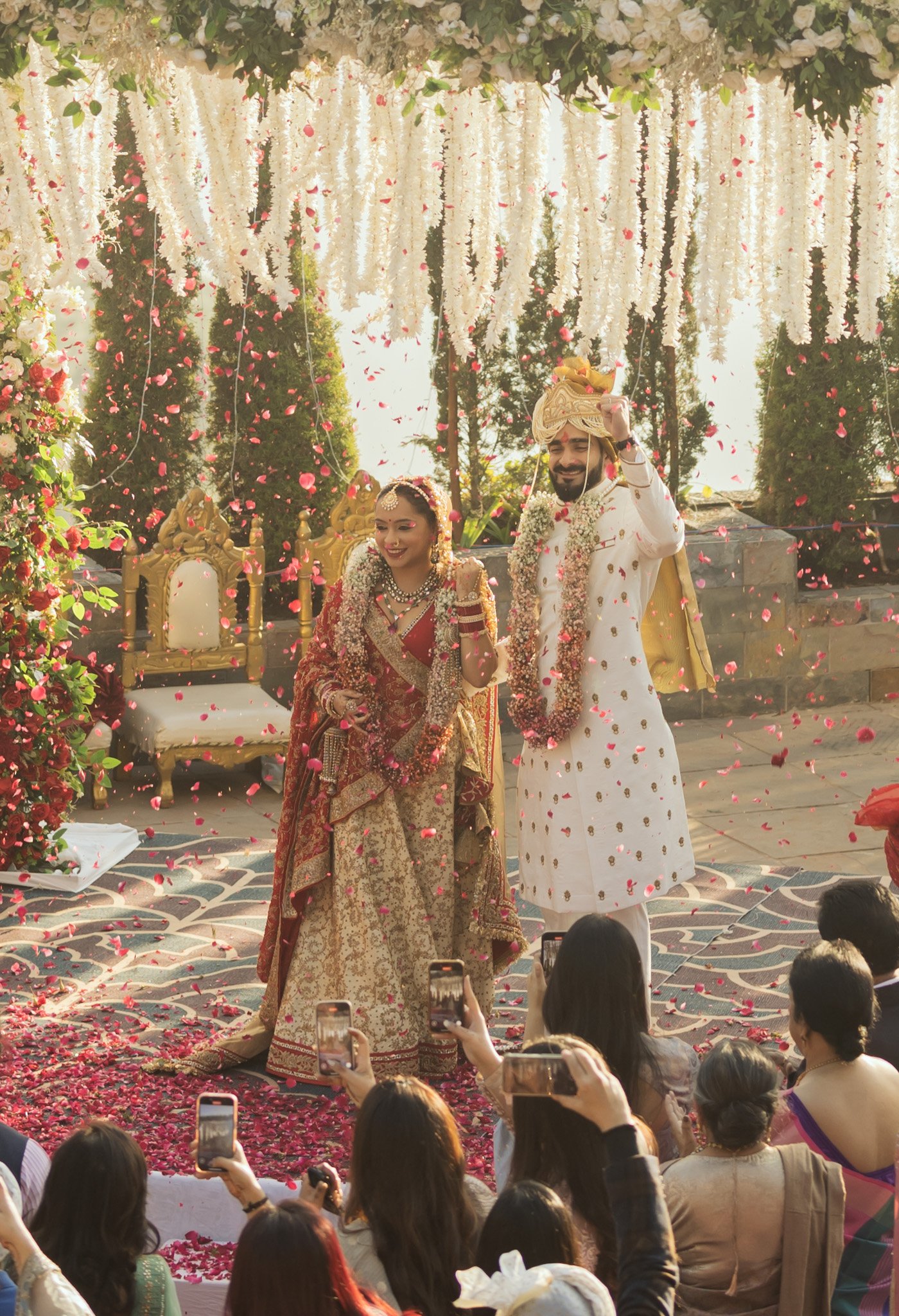 Bride and groom in traditional Indian wedding attire standing on a stage decorated with flowers, surrounded by guests taking photos, during a wedding ceremony.
