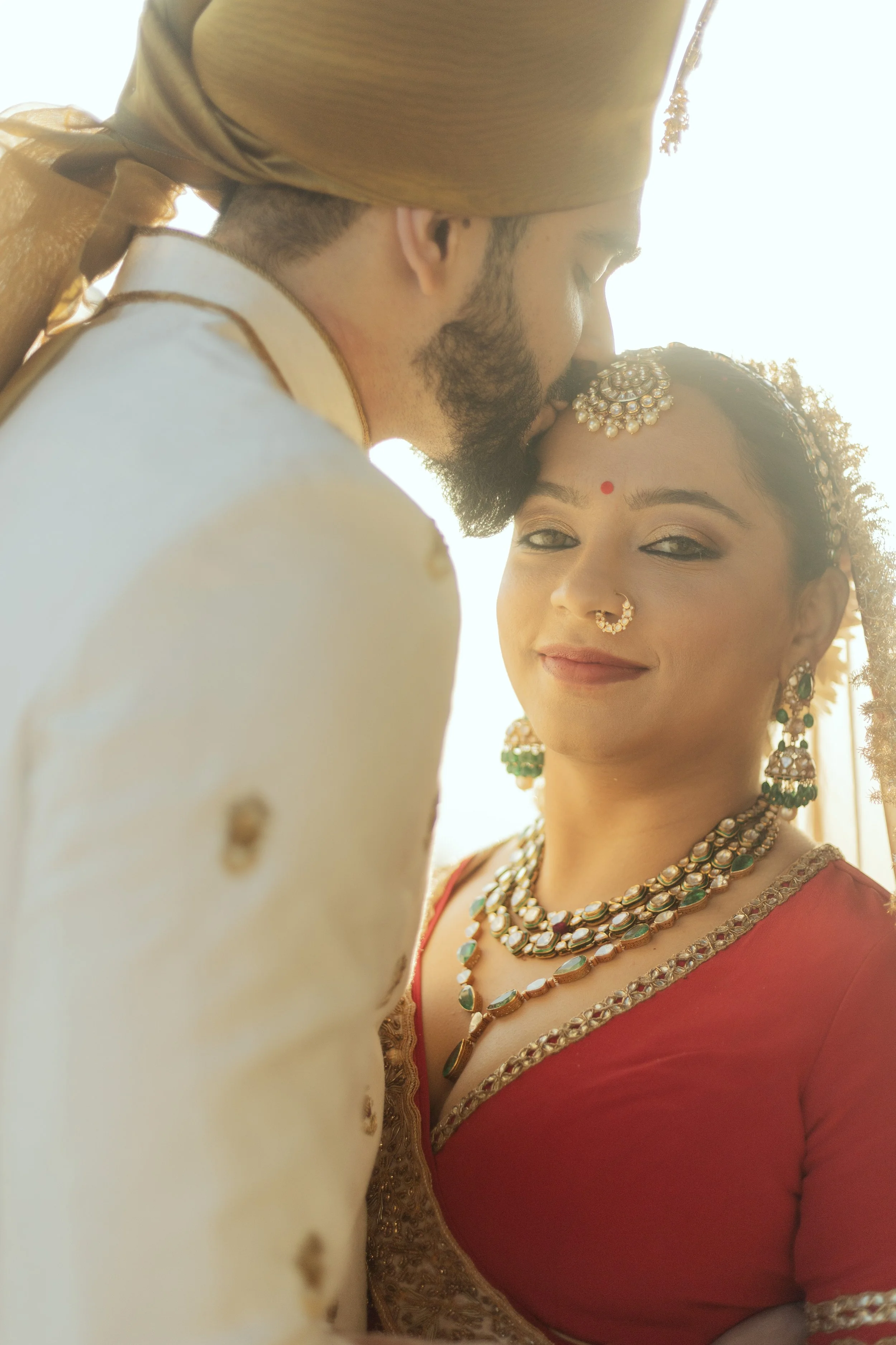 A close-up photo of a bride and groom in traditional Indian attire, sharing an intimate moment with the groom kissing the bride's forehead, both wearing ornate jewelry.