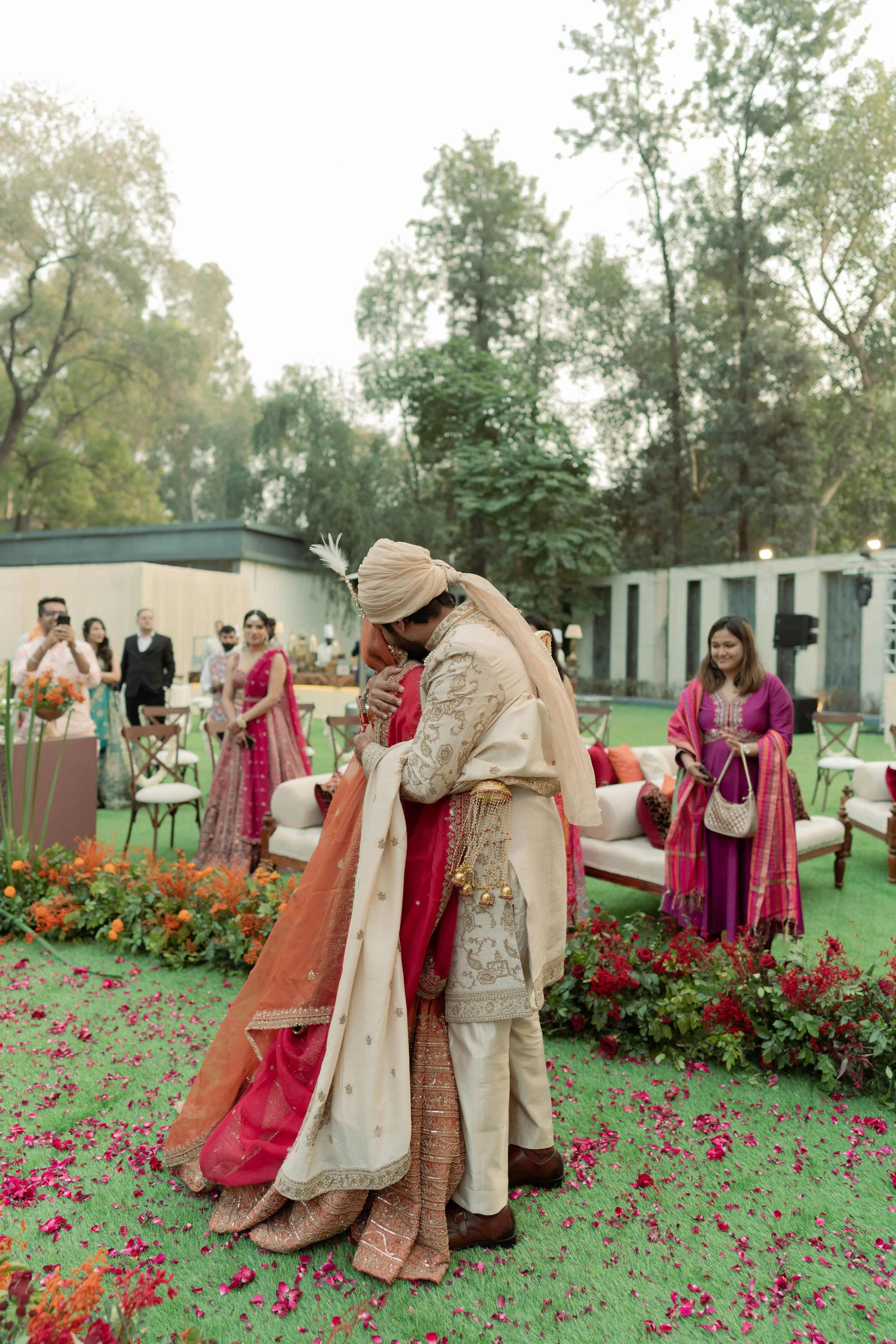 A groom and bride embracing during a wedding ceremony outdoors, with guests in traditional attire in the background.
