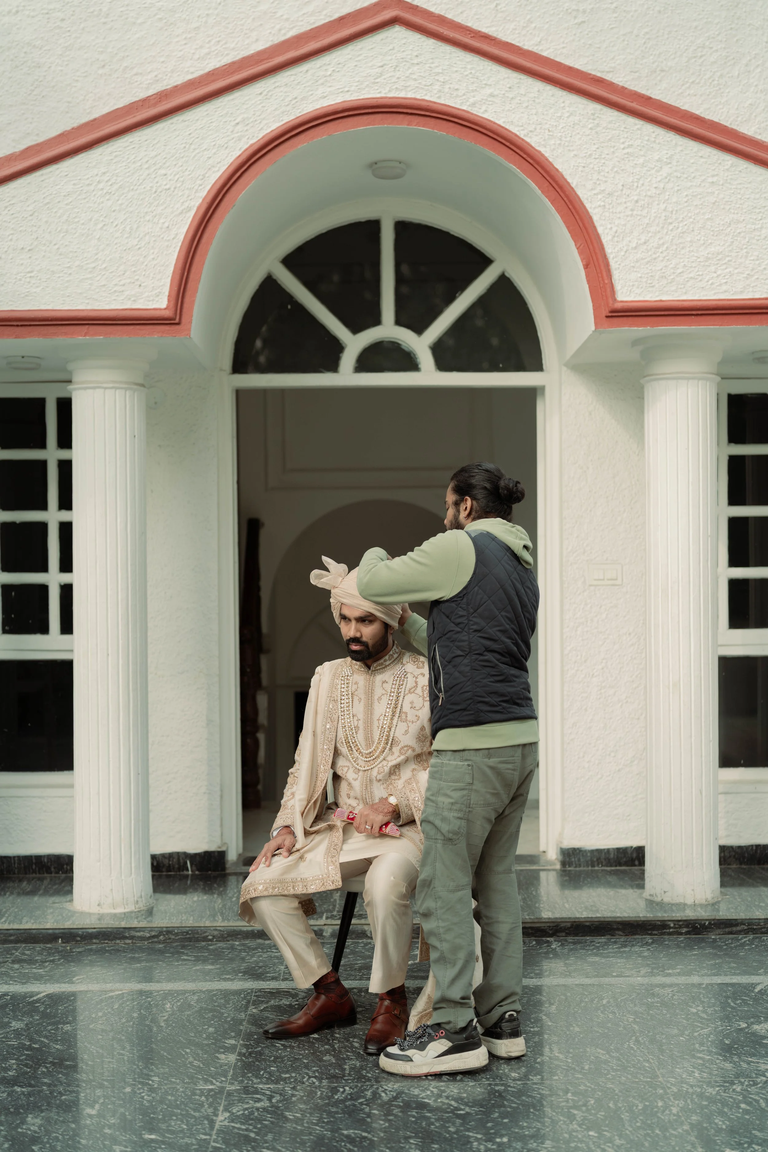 A man dressed in traditional Indian wedding attire sits on a chair while another man adjusts his turban outside a house with white columns and arched windows.