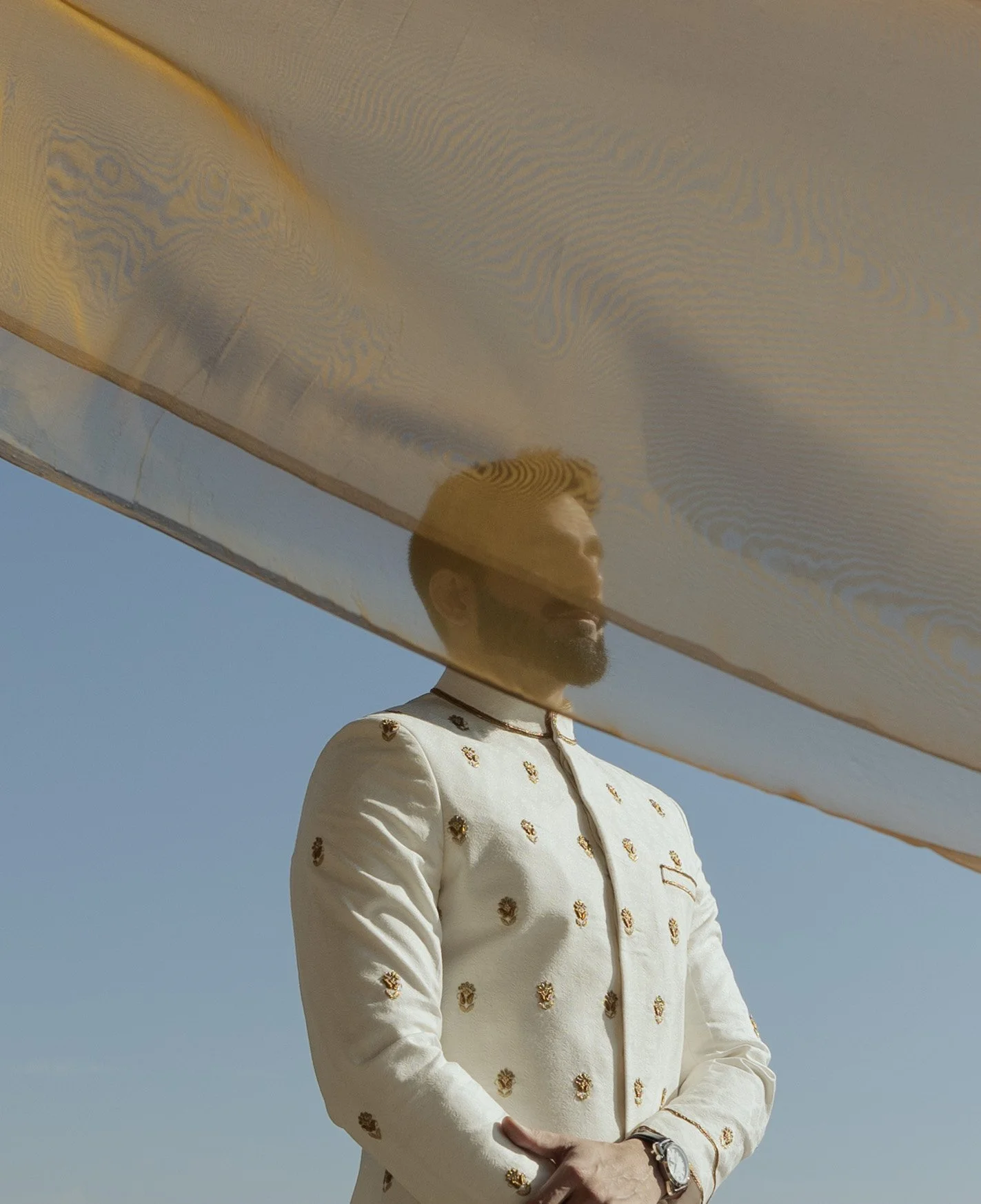 A man with a beard and short hair, dressed in a white traditional Indian sherwani with gold embroidery, stands outdoors underneath a large reflective metallic structure. The sky is clear and blue.