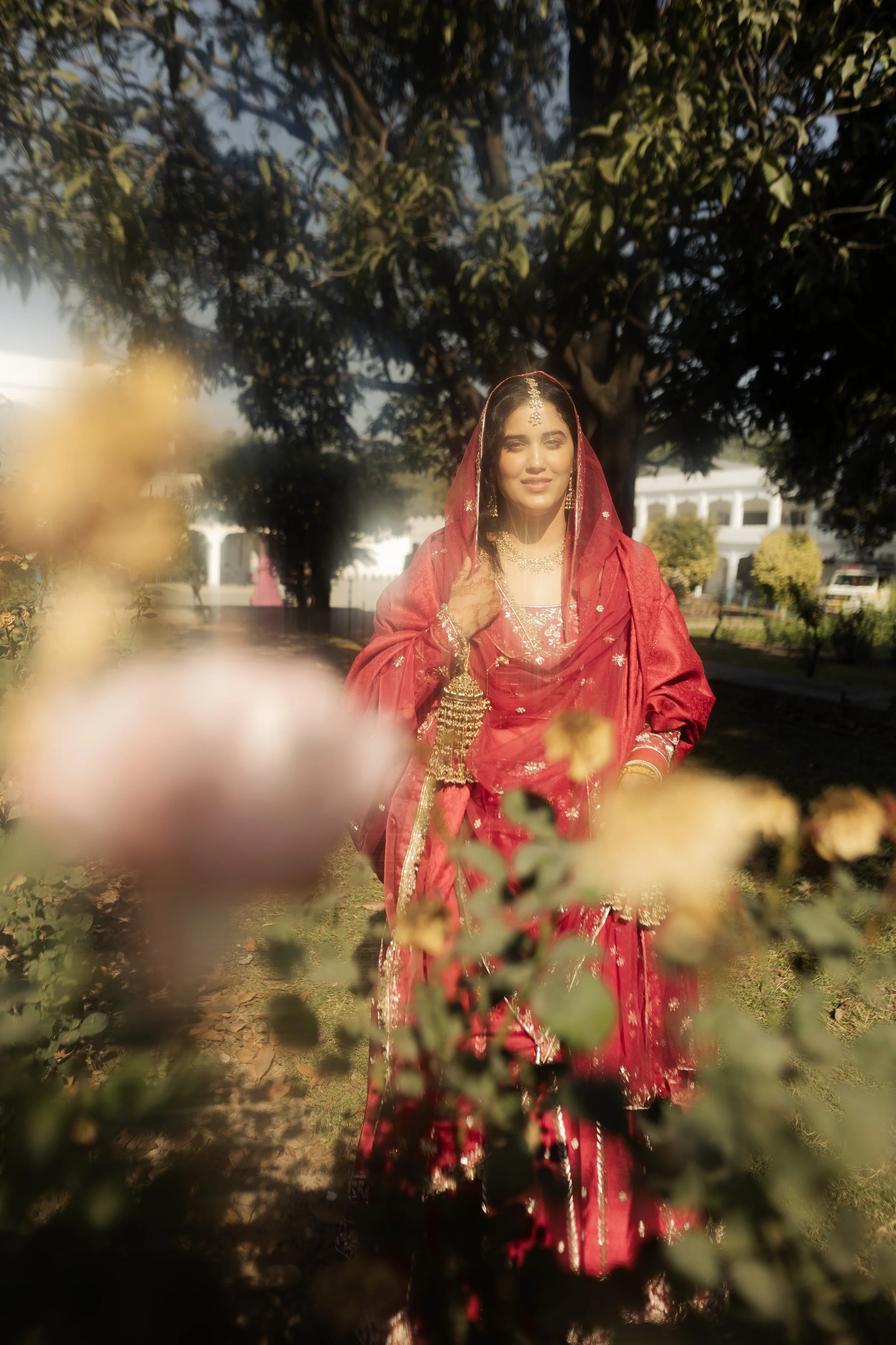 A woman dressed in traditional red Indian attire, adorned with jewelry and a veil, standing outdoors with trees and houses in the background.