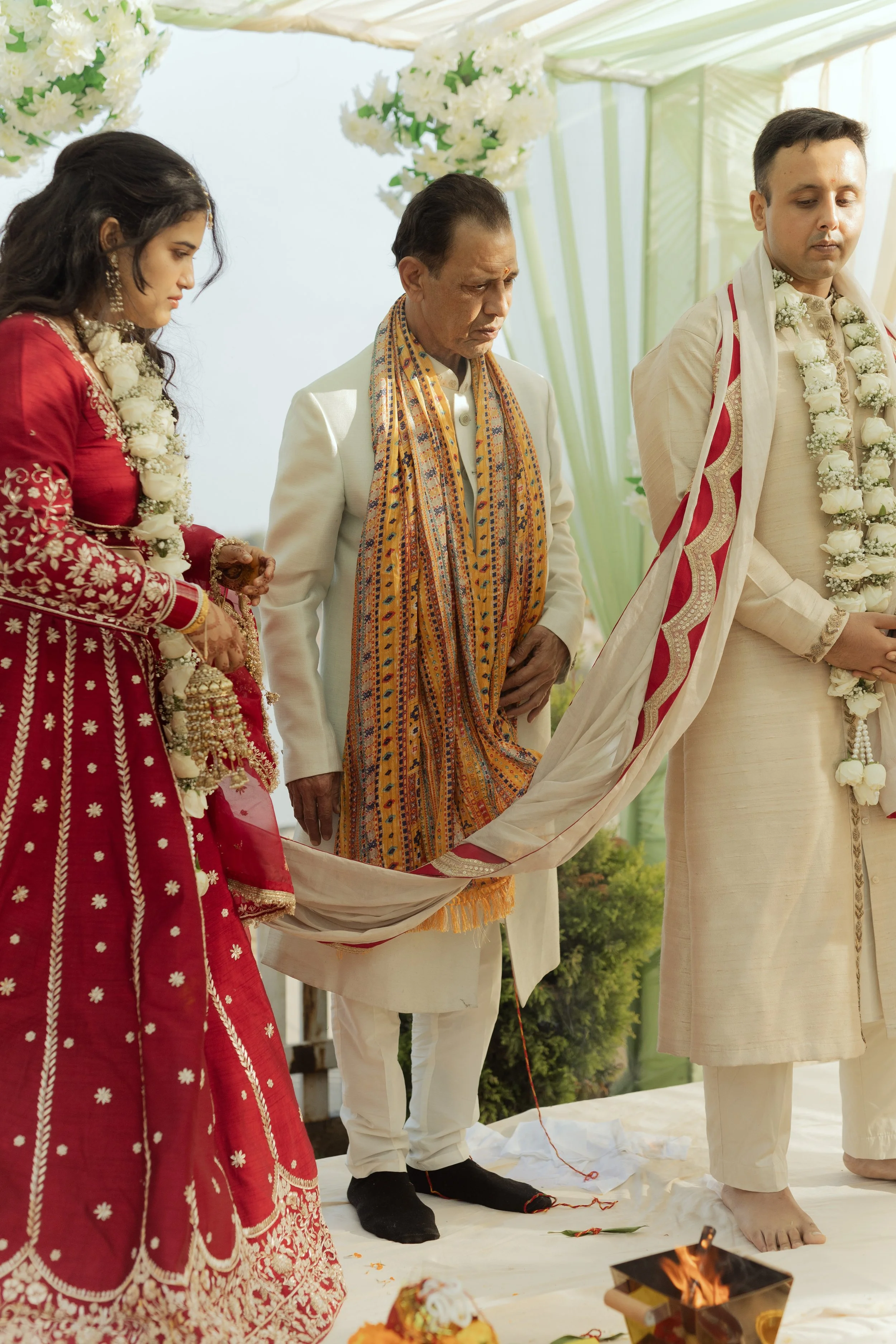Three people participating in a traditional Indian wedding ceremony. The bride is wearing a red embroidered dress, the groom is in cream attire with a floral garland, and an older man in white with a colorful scarf is standing between them with bowed