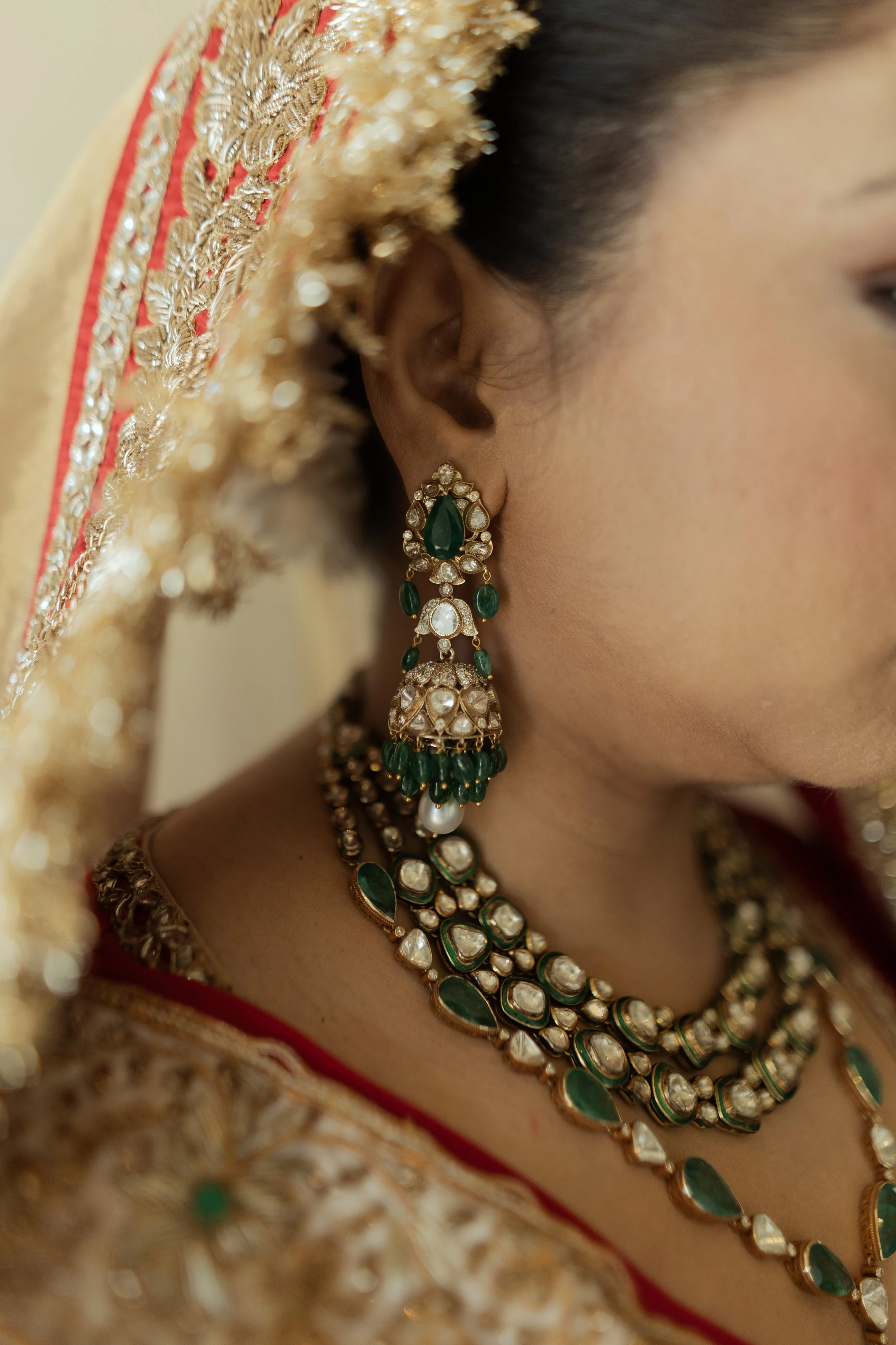 Close-up of a woman wearing traditional Indian jewelry, including large earrings and layered necklaces, with her hair covered by a richly embroidered dupatta or veil.