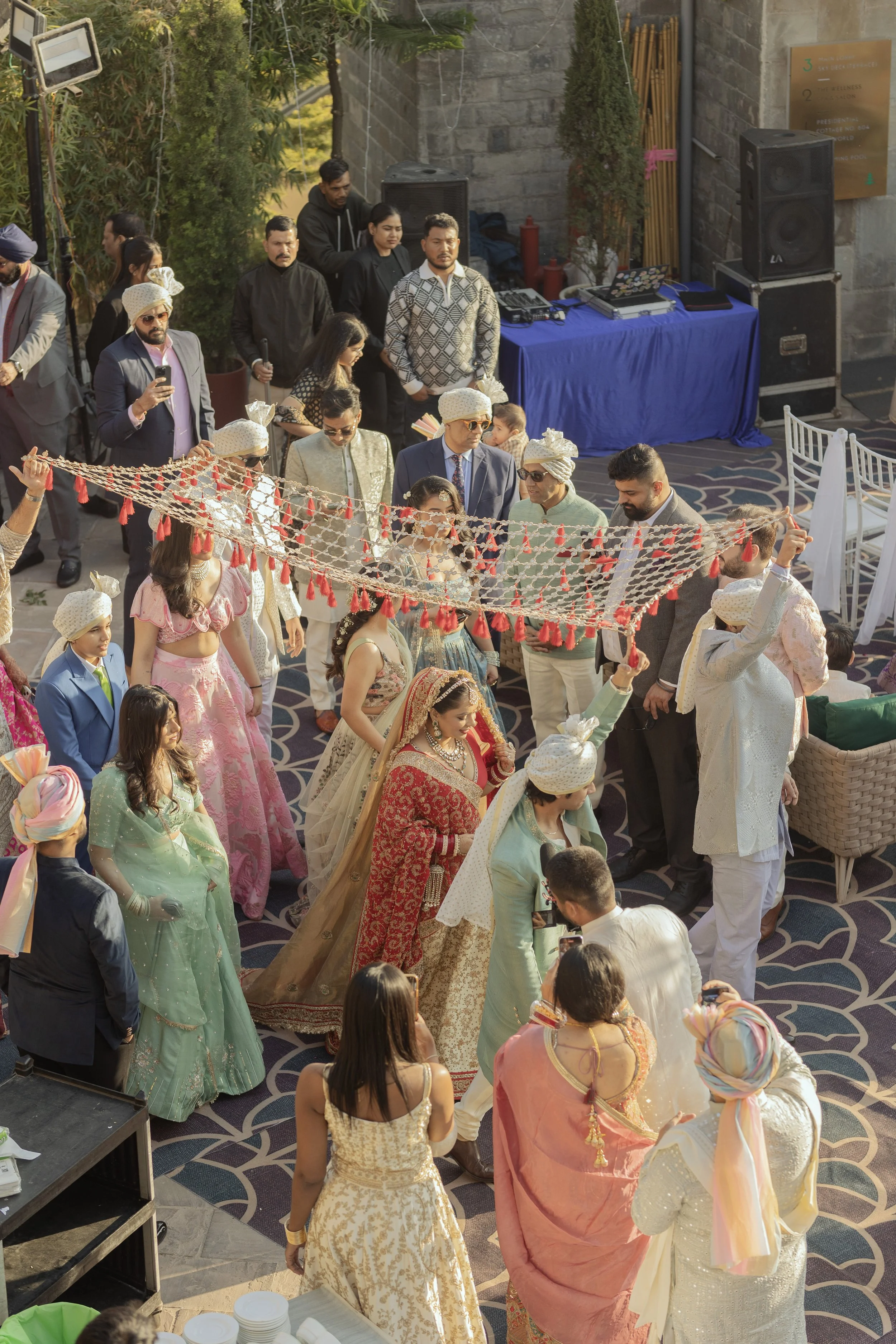Group of people participating in a traditional Indian wedding ceremony, holding a decorative cloth, dressed in colorful sarees, suits, and turbans, with a DJ setup in the background.