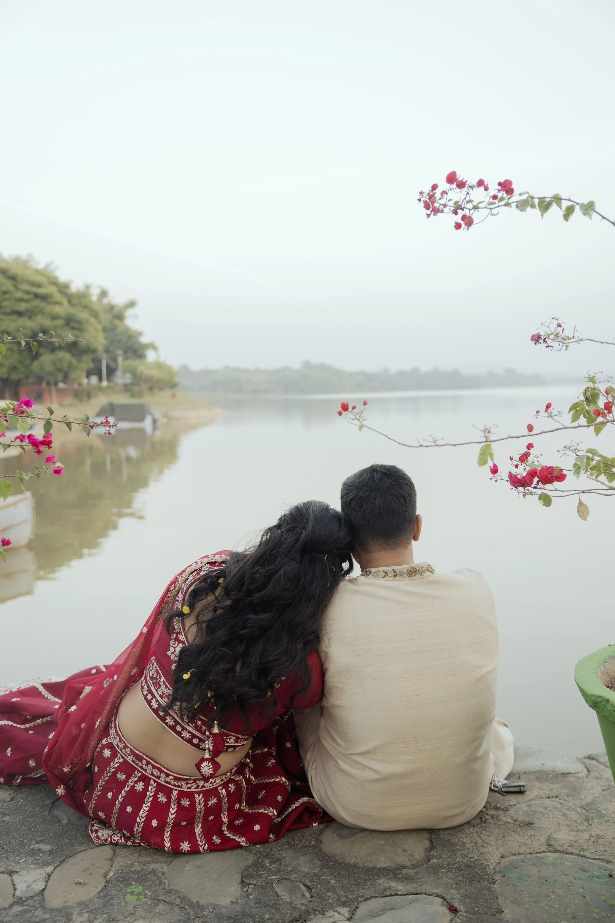 A couple sitting on a stone surface by the water, with the woman resting her head on the man's shoulder, overlooking a calm lake with trees and flowering branches in the foreground.