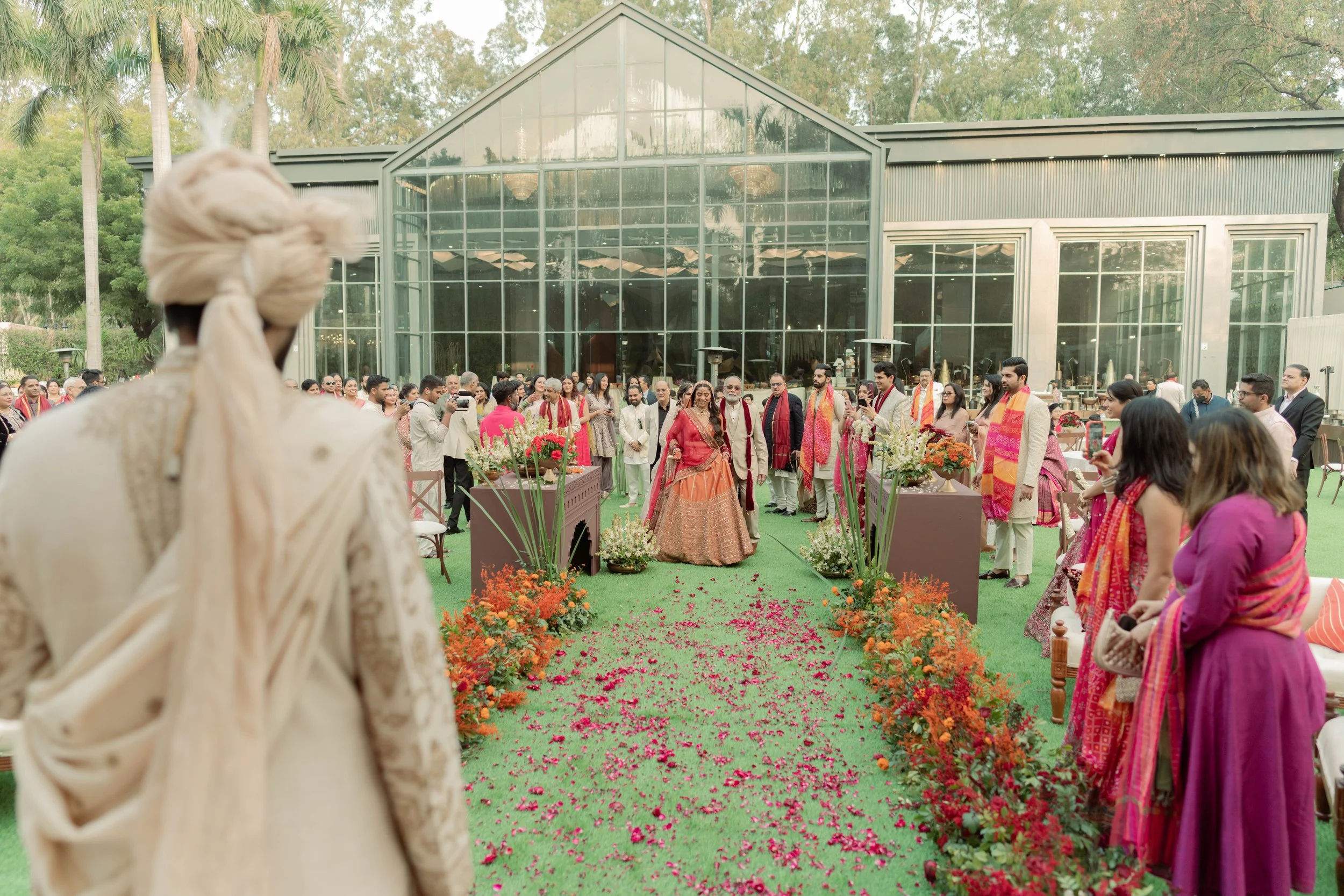 Indian wedding ceremony outdoors with guests in traditional attire, floral decorations, and a brick building with glass windows in the background.