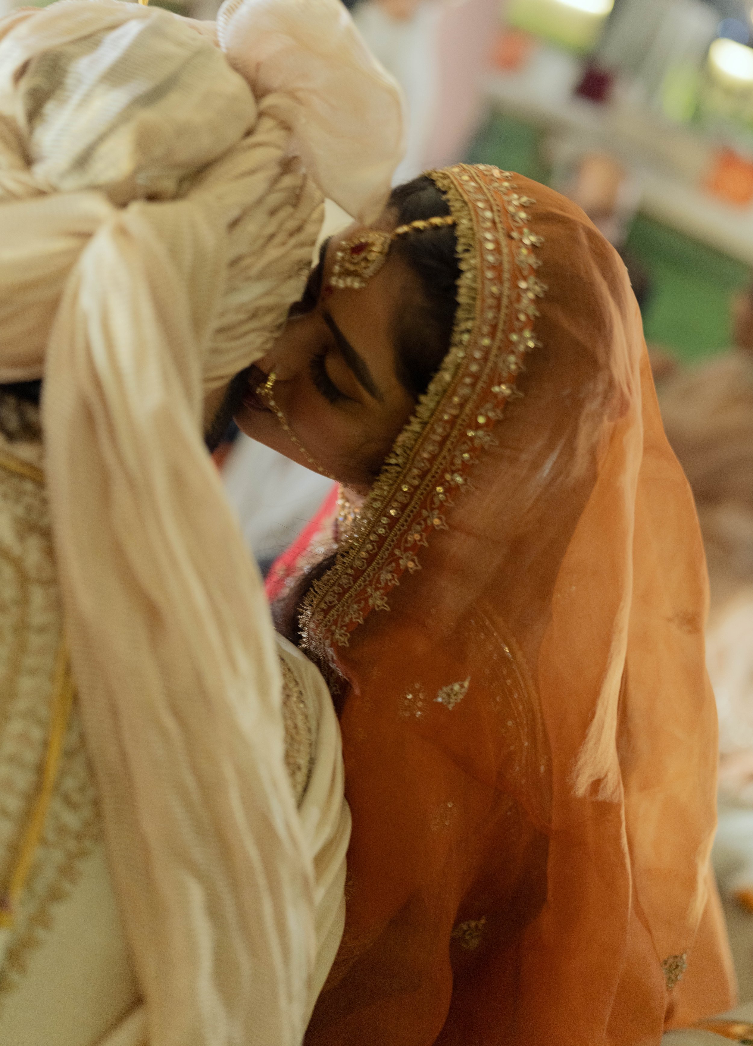 A bride and groom share a kiss during a wedding ceremony, with the bride wearing a traditional orange saree and intricate jewelry.