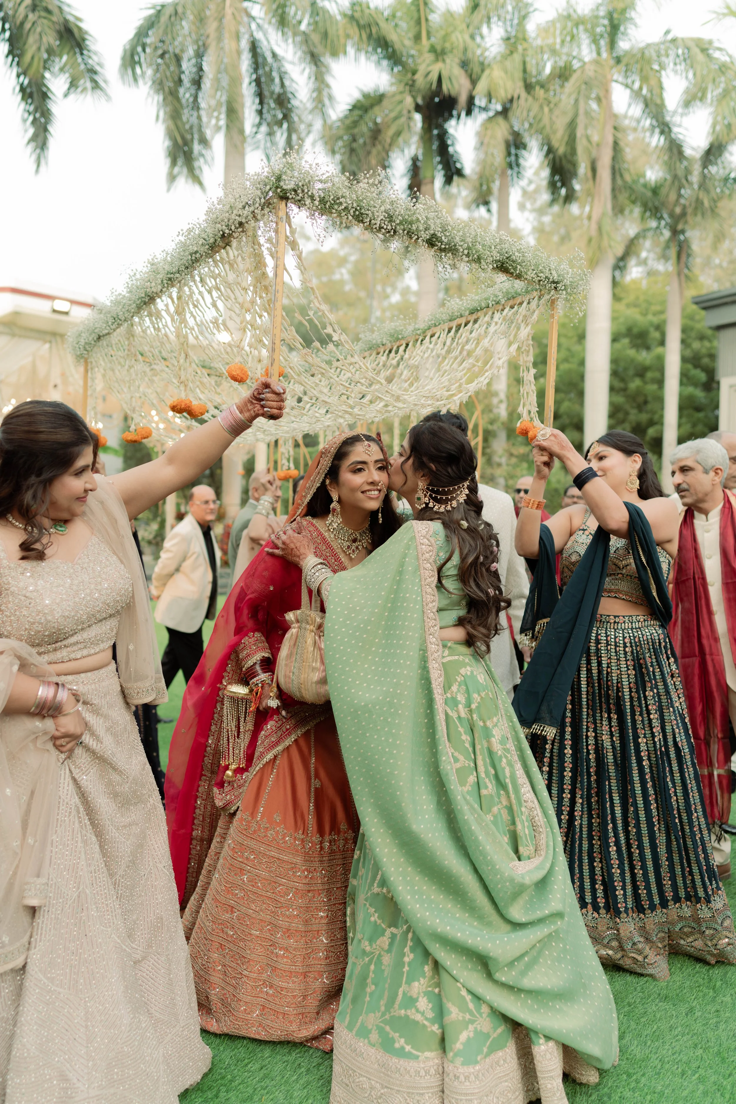 Indian wedding celebration with women dressed in colorful traditional attire under a decorated canopy, surrounded by lush greenery and tall trees.