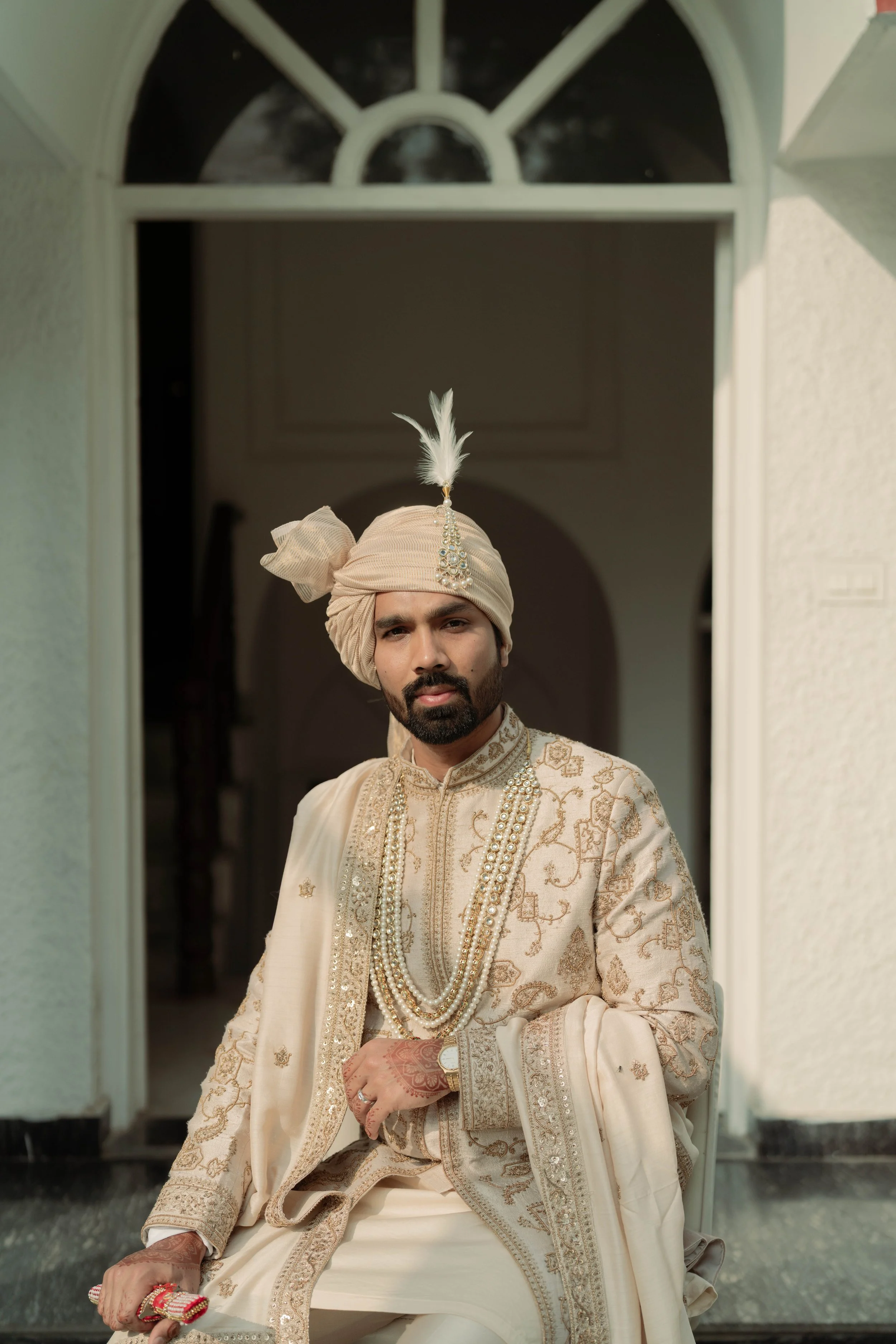 A man dressed in traditional Indian wedding attire, wearing a cream-colored sherwani with intricate embroidery, layered pearl necklaces, a cream turban with a feather and jewel ornament, sitting in front of an arched doorway.