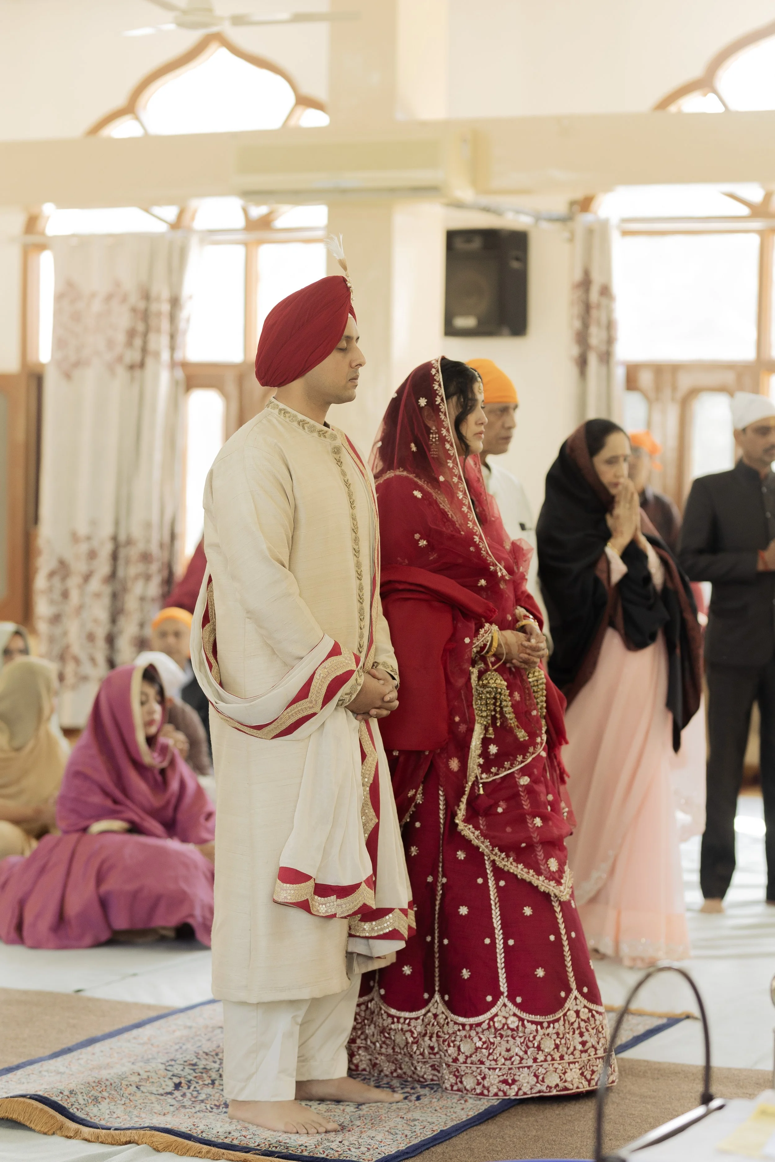 Indian wedding ceremony with a groom and bride standing in prayer, surrounded by family members in traditional attire inside a decorated hall.