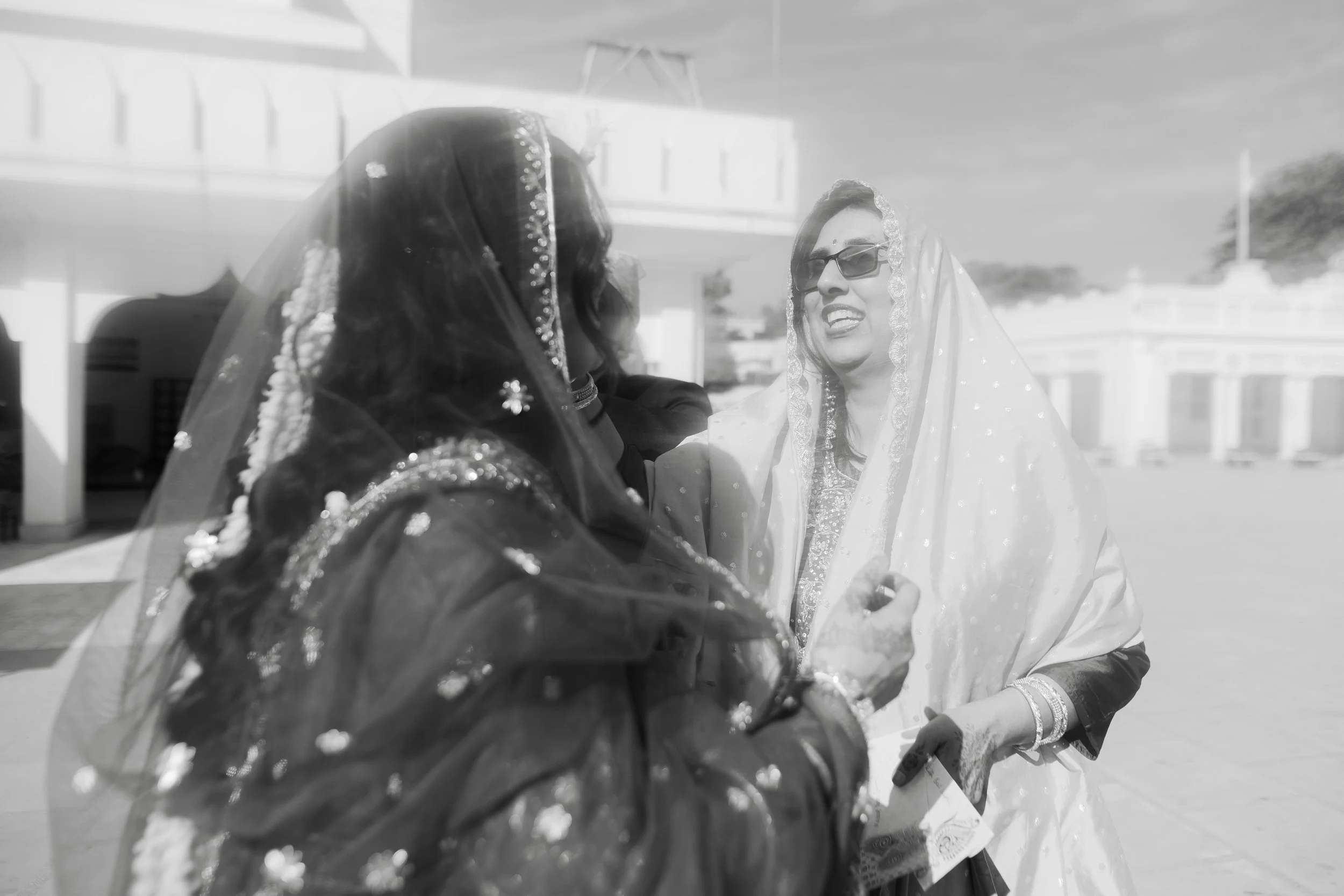 Two women wearing traditional Indian attire and sarees, smiling and engaging in a conversation outdoors during daylight.