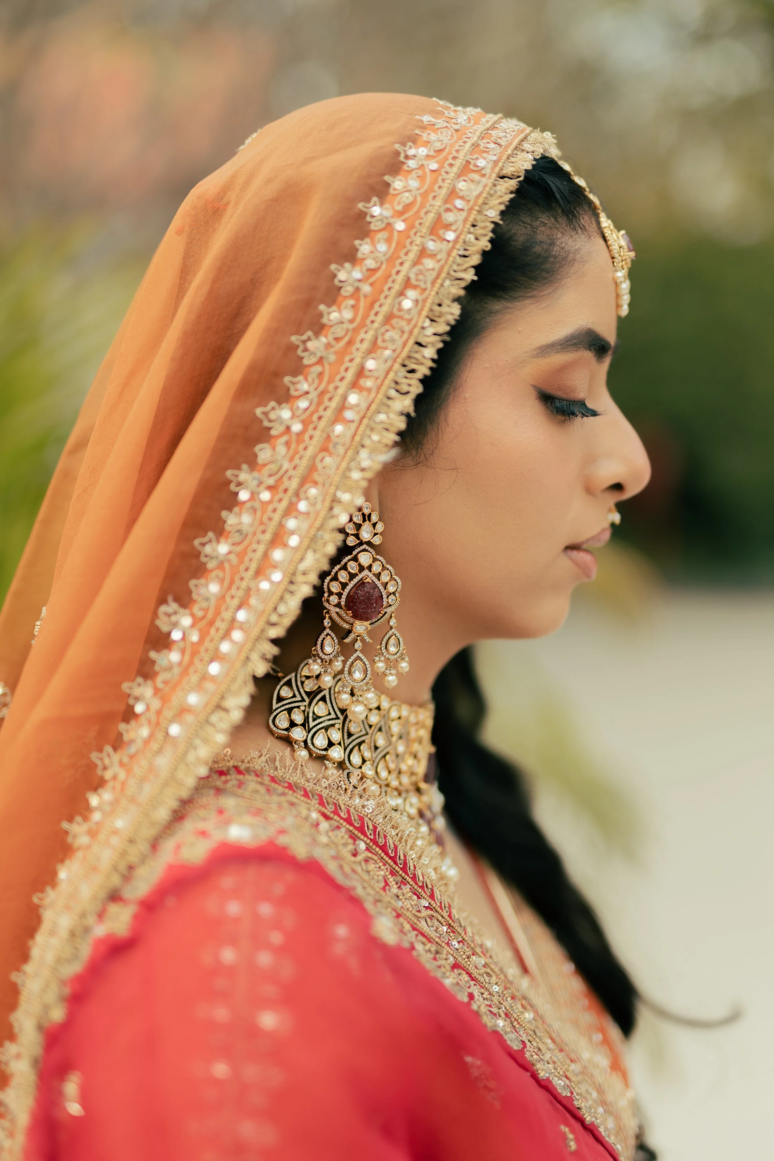 Close-up of a woman in traditional Indian attire with a peach-colored dupatta adorned with floral embroidery and sequins, earrings, and jewelry, with her eyes closed, outdoors with a blurred background.
