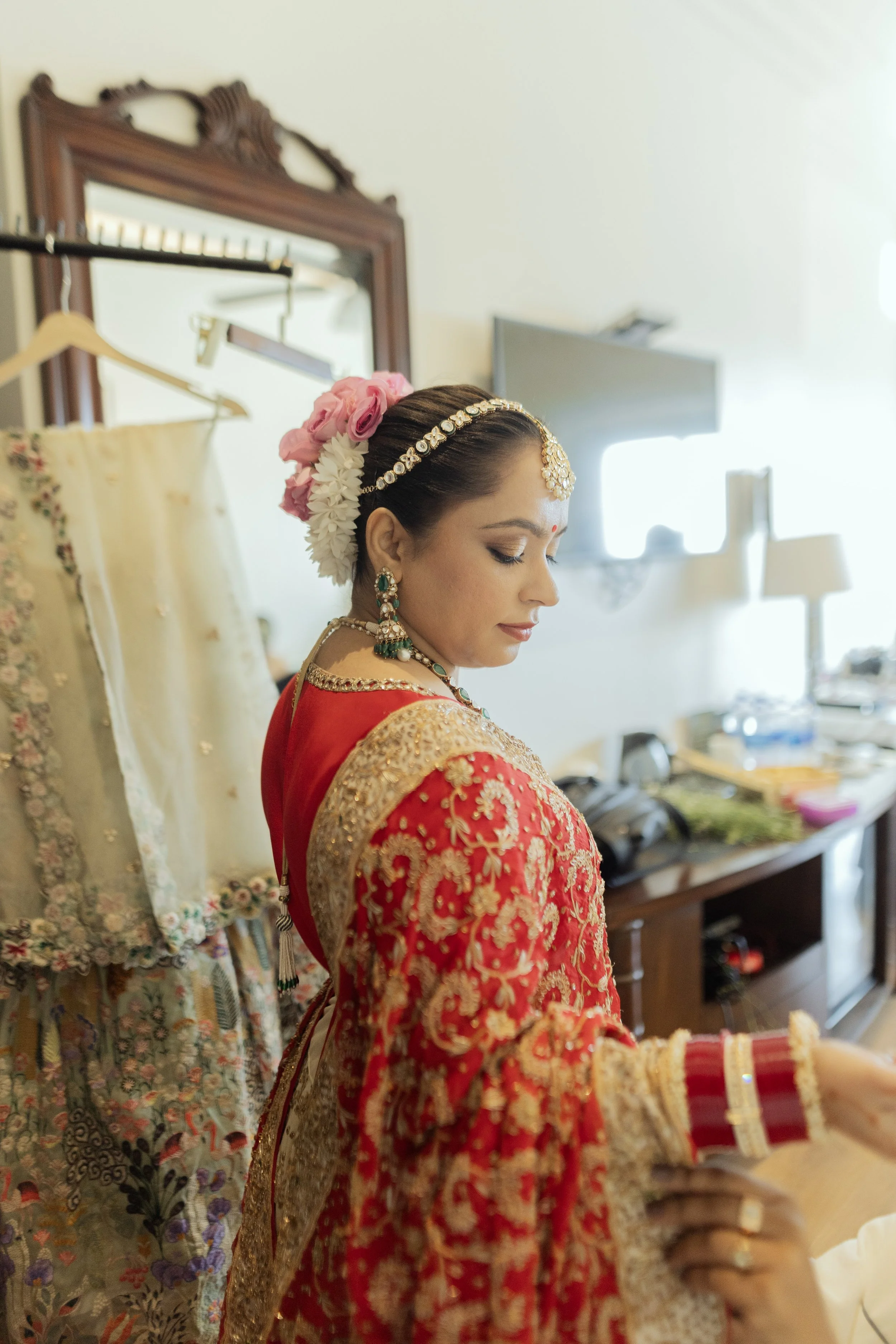 A bride in traditional Indian attire is preparing for her wedding, wearing a red and gold embroidered saree, adorned with jewelry, including earrings, necklaces, and a forehead ornament, with flowers in her hair.