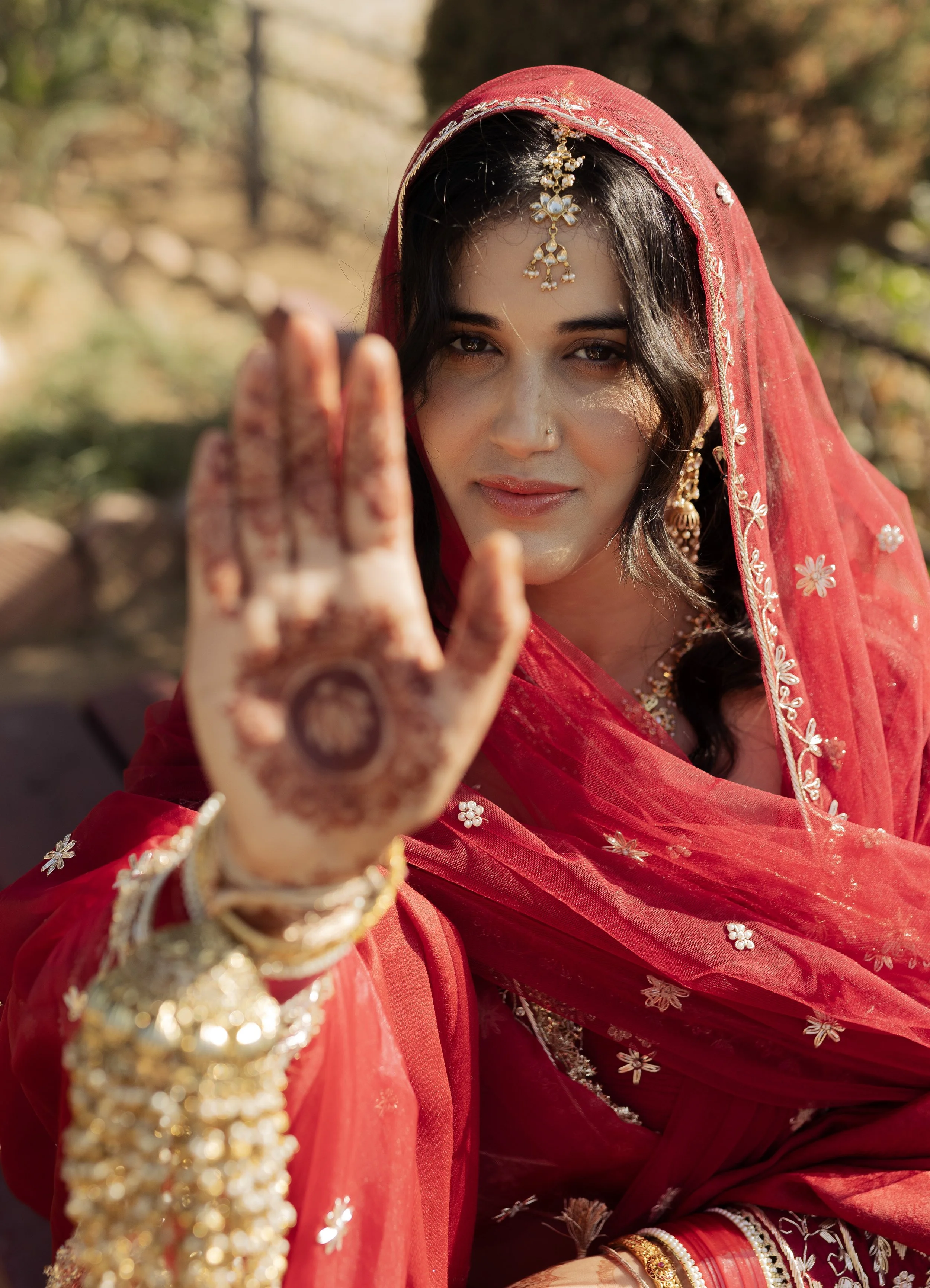 A woman dressed in traditional Indian attire, wearing a red saree and jewelry, holding her hand up with henna designs on her palm, outdoors with trees in the background.