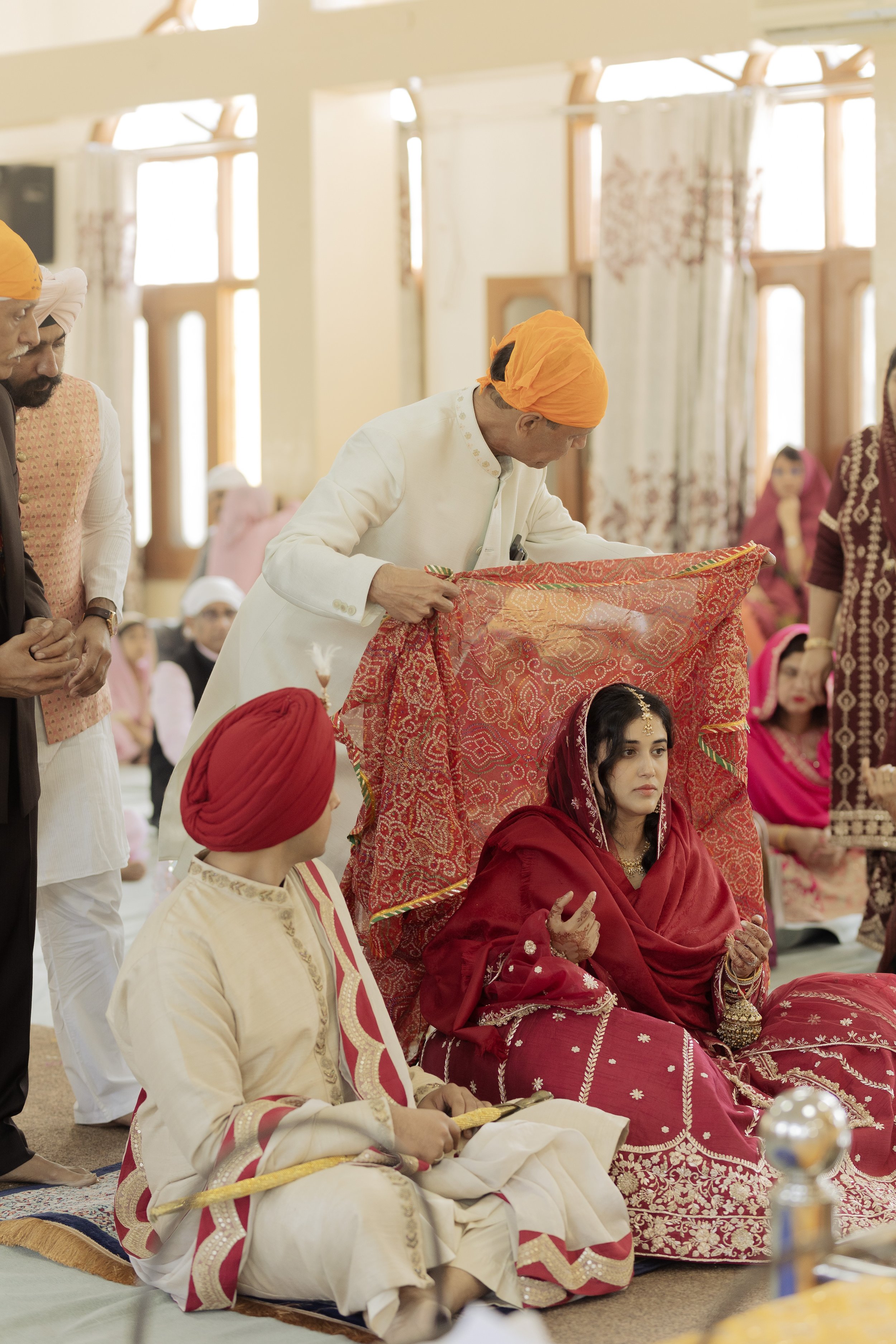A traditional Indian wedding ceremony with the bride and groom dressed in elaborate attire, seated on the floor, while officiants perform rituals under a canopy with vibrant red and gold fabric.