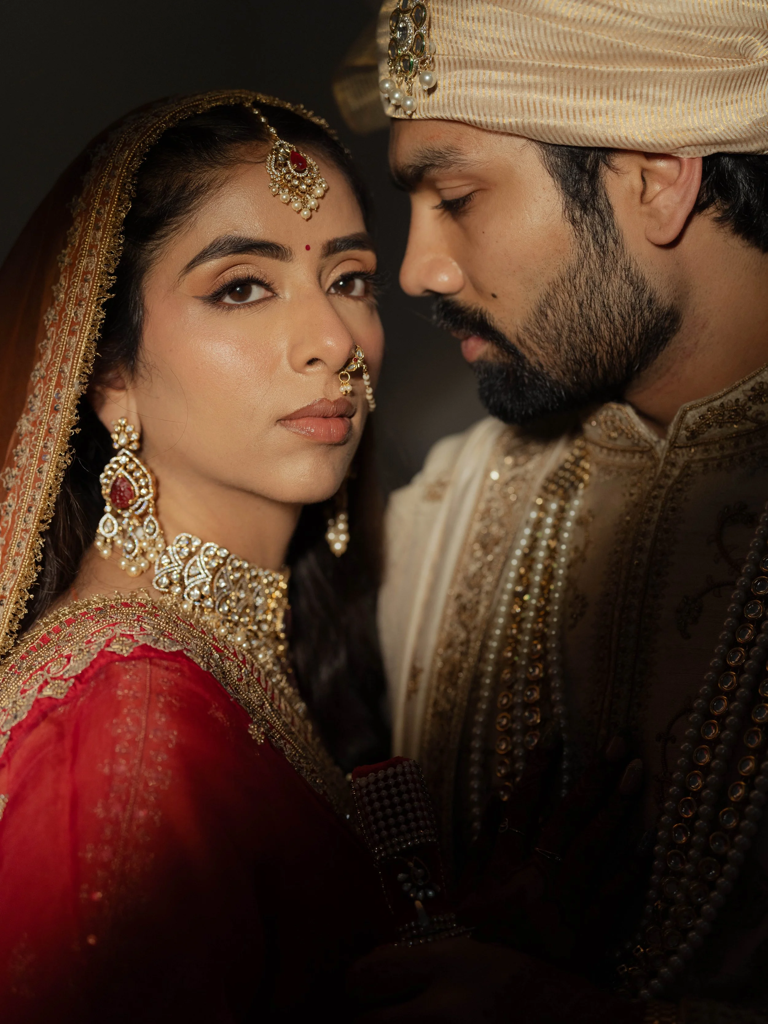 Close-up of a bride and groom in traditional Indian wedding attire, gazing at each other, bride wearing elaborate jewelry and red dress, groom in cream-colored sherwani and turban.