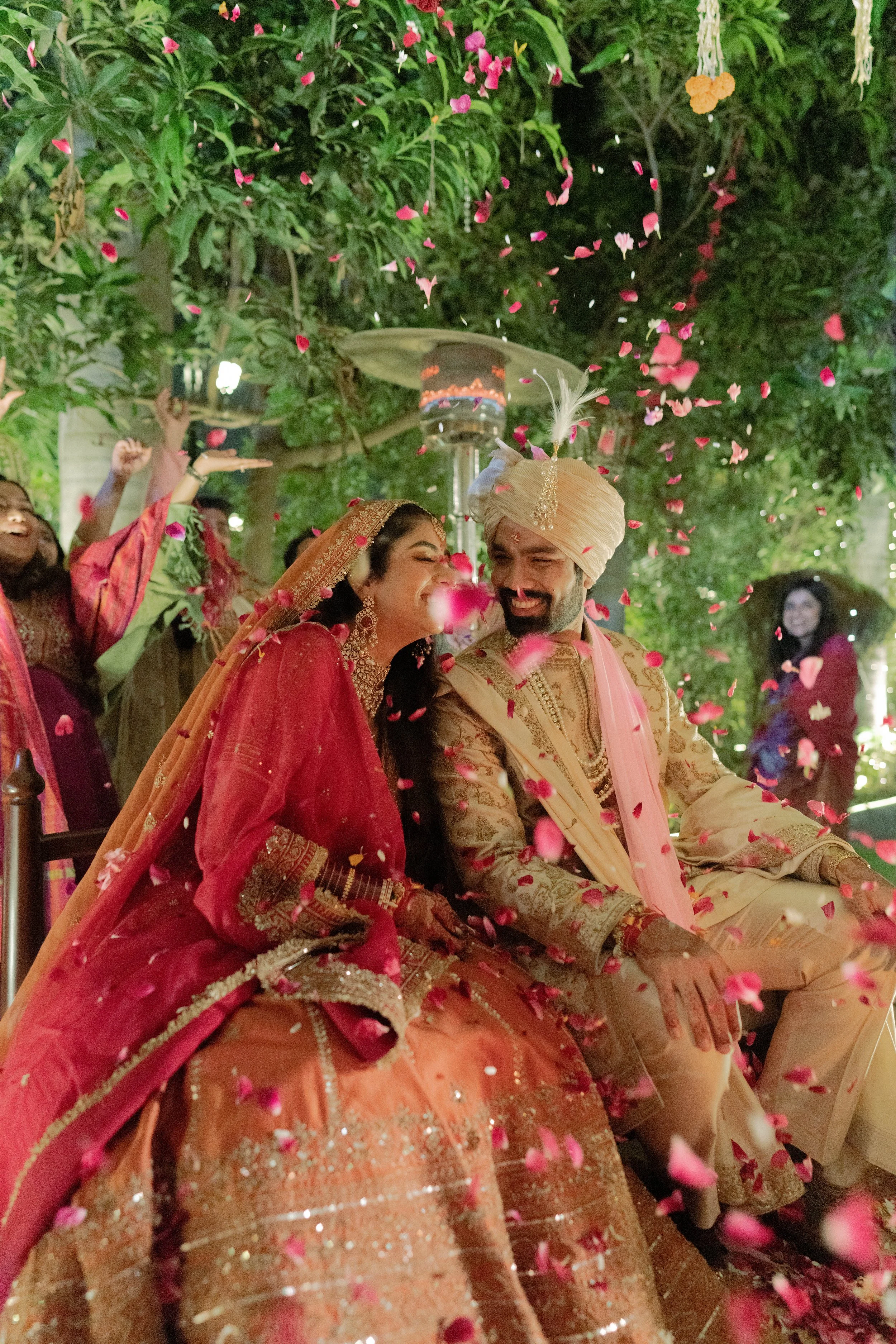An Indian bride and groom in traditional wedding attire sitting together during their wedding celebration, surrounded by pink flower petals and friends celebrating around them.