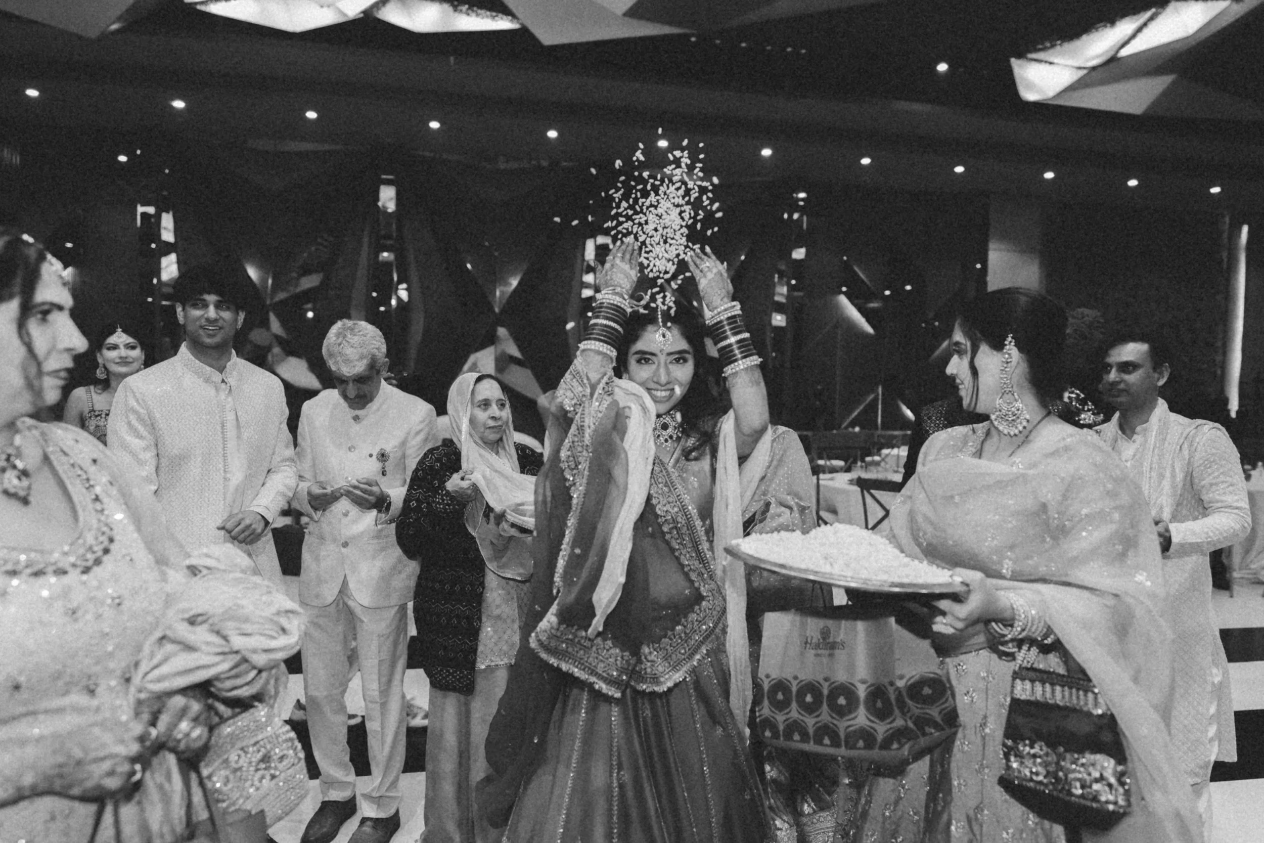 A group of people at an Indian wedding celebrating a bride who is sprinkling rice overhead. The bride is smiling and wearing traditional jewelry and attire. Several women and men in traditional Indian clothing are present, some holding trays of rice,