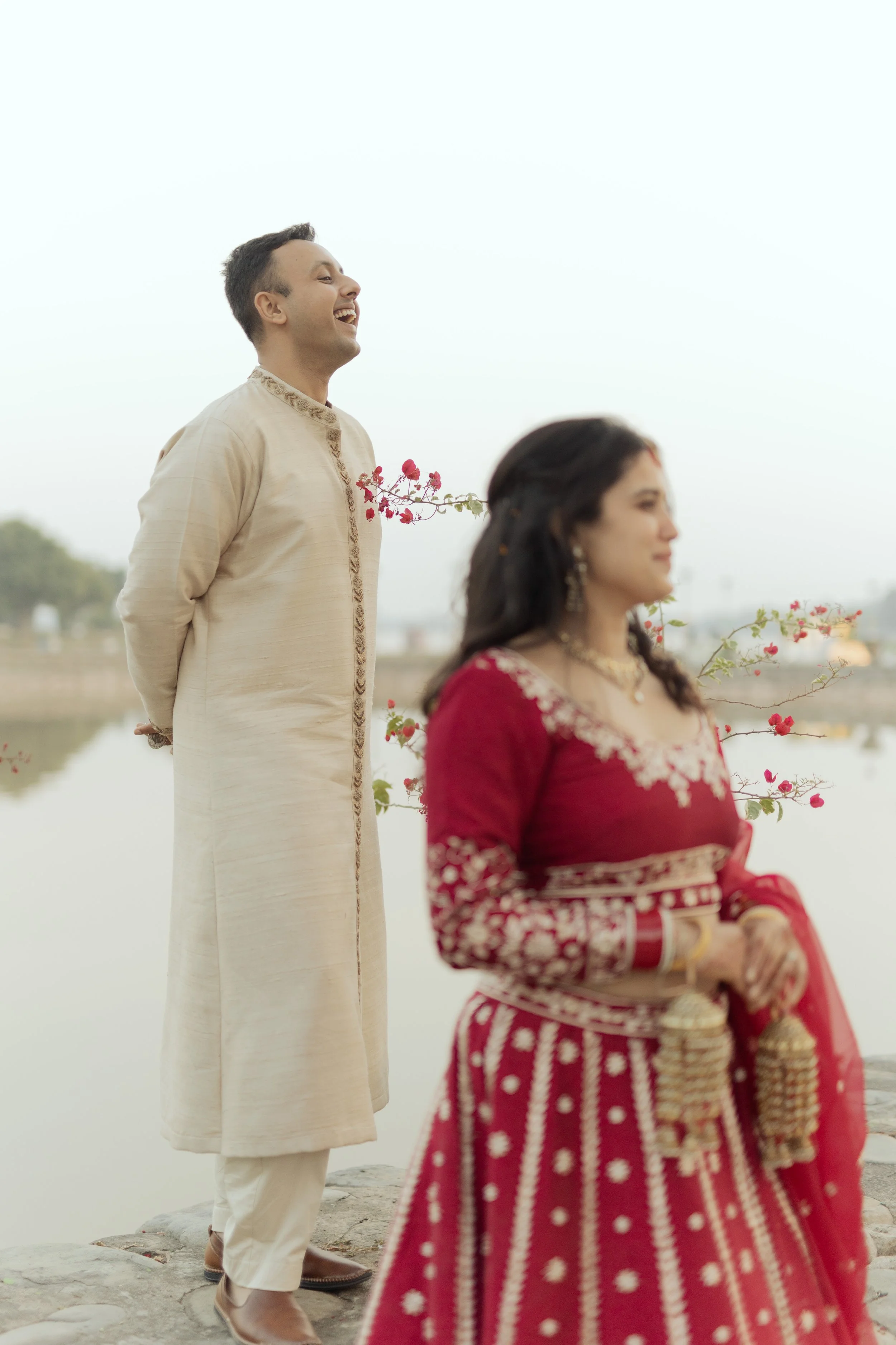 A couple in traditional Indian attire standing outdoors near a body of water, with the man smiling and looking up, and the woman standing with her eyes closed holding red and gold accessories.