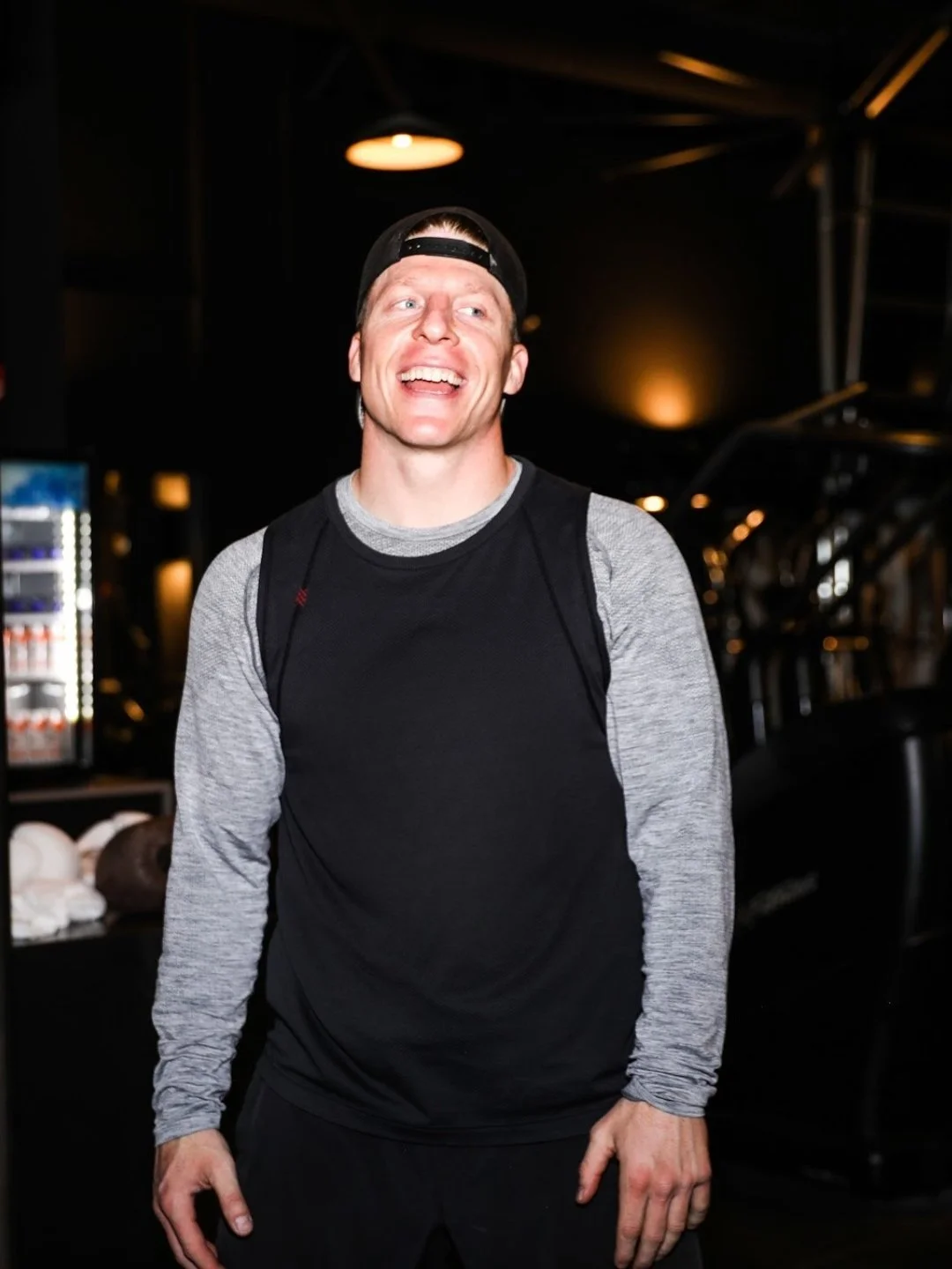 A man smiling and talking in a gym, wearing a black and gray long sleeve athletic shirt and a black backward cap.