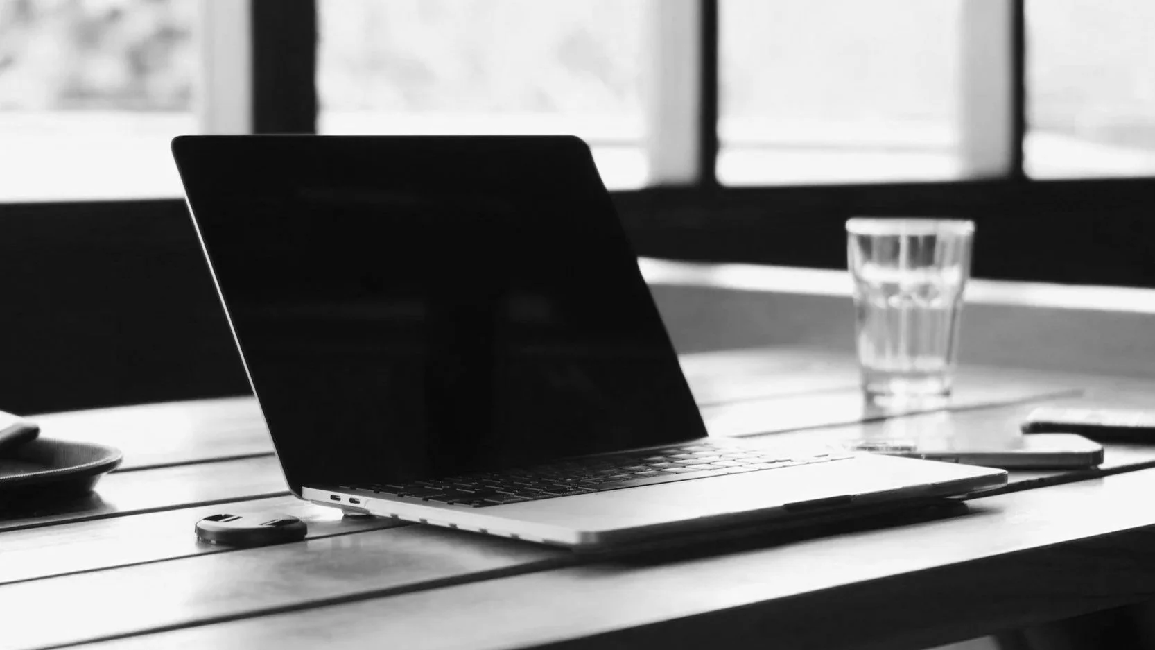 A black and white photo of a laptop, a glass of water, a mouse, and a small device on a wooden table with sunlight coming through windows in the background