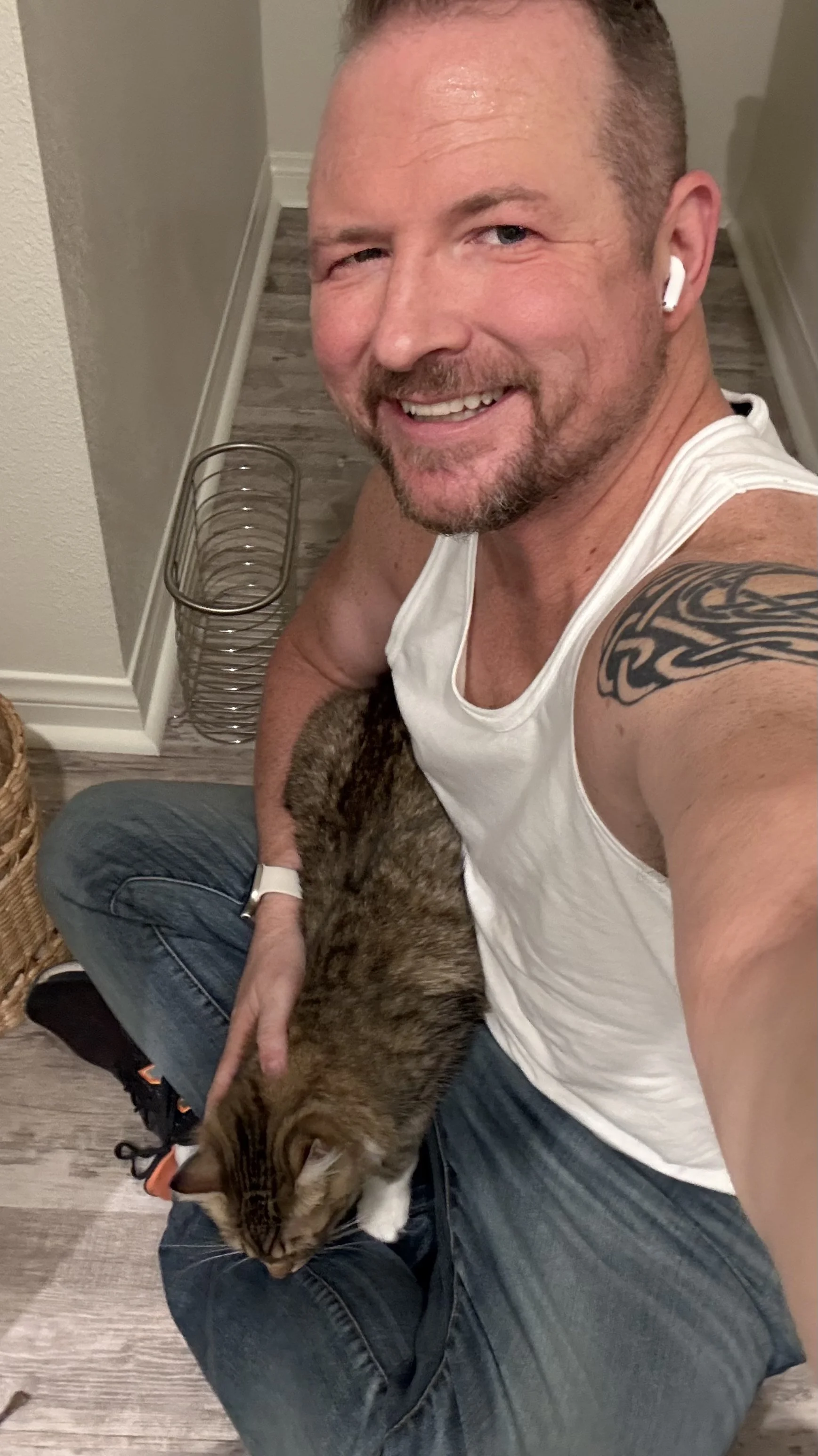 A smiling man with tattoos and AirPods taking a selfie while holding a cat on his lap.