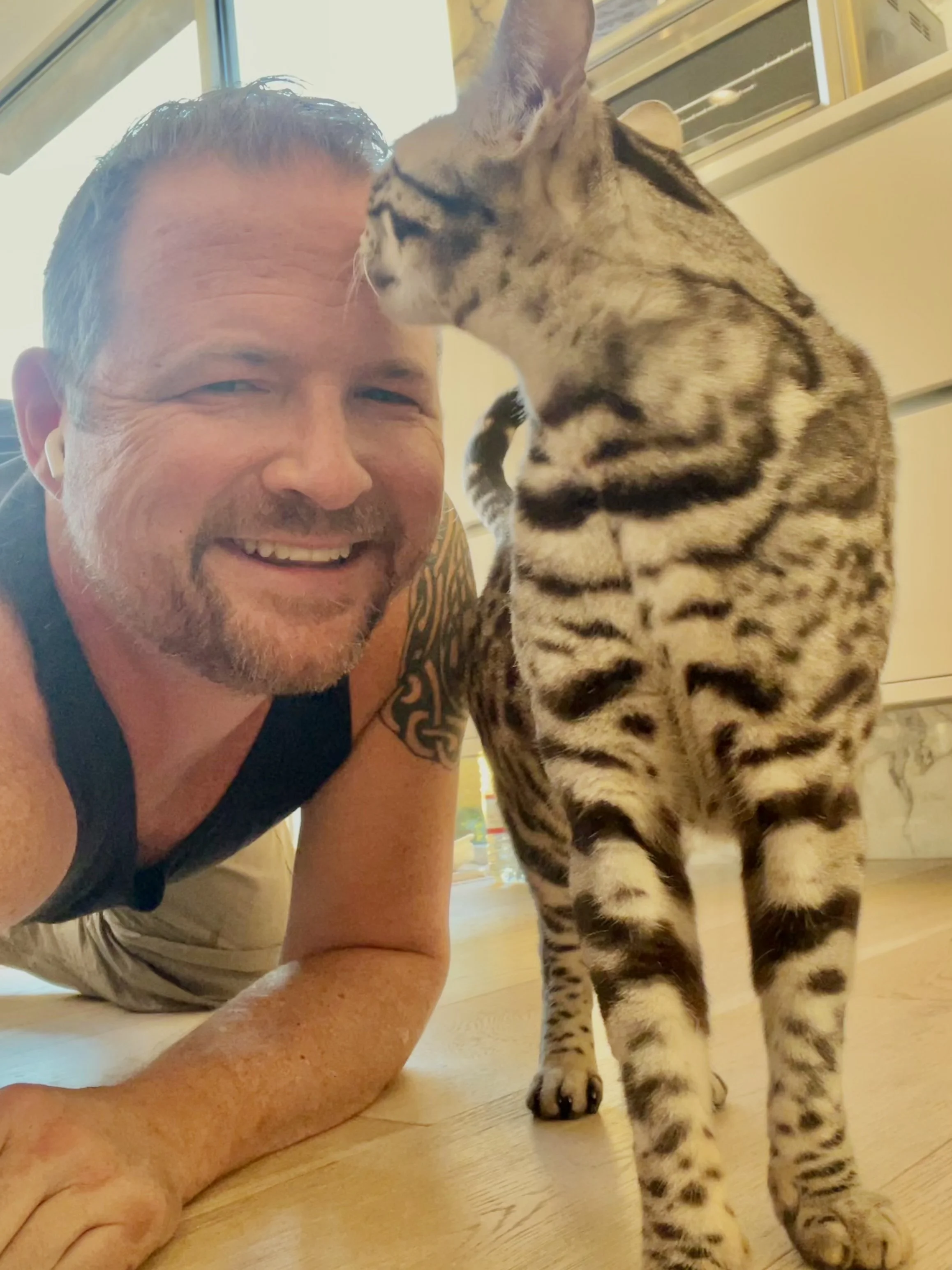 A man lying on the floor smiling with a Bengal cat standing next to him. The man has short hair, a beard, and tattoos on his arm. The cat has a spotted coat and is nuzzling his forehead.