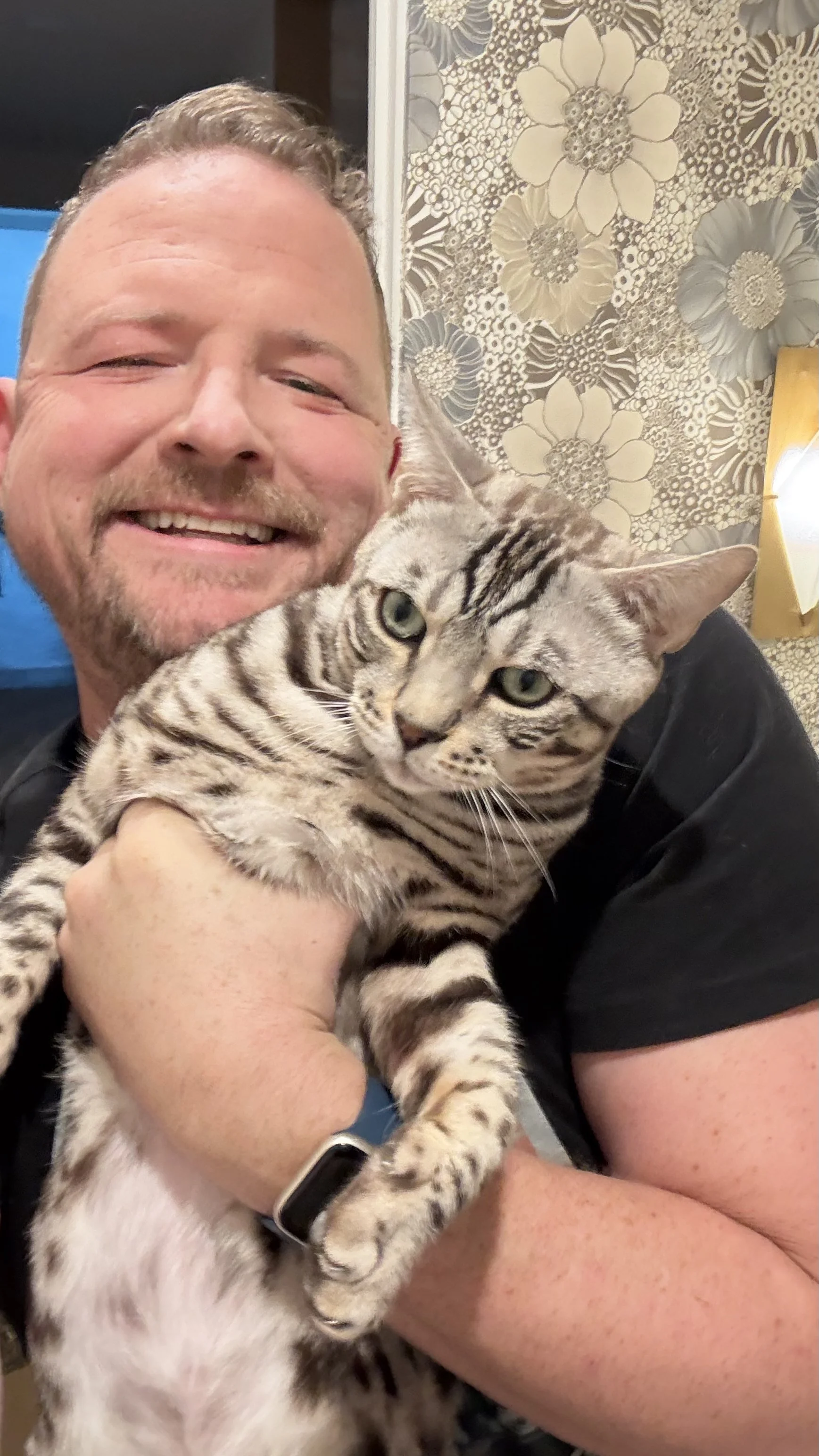 A smiling man holds a Bengal cat indoors, with a ornate floral wallpaper and a yellow light art piece in the background.