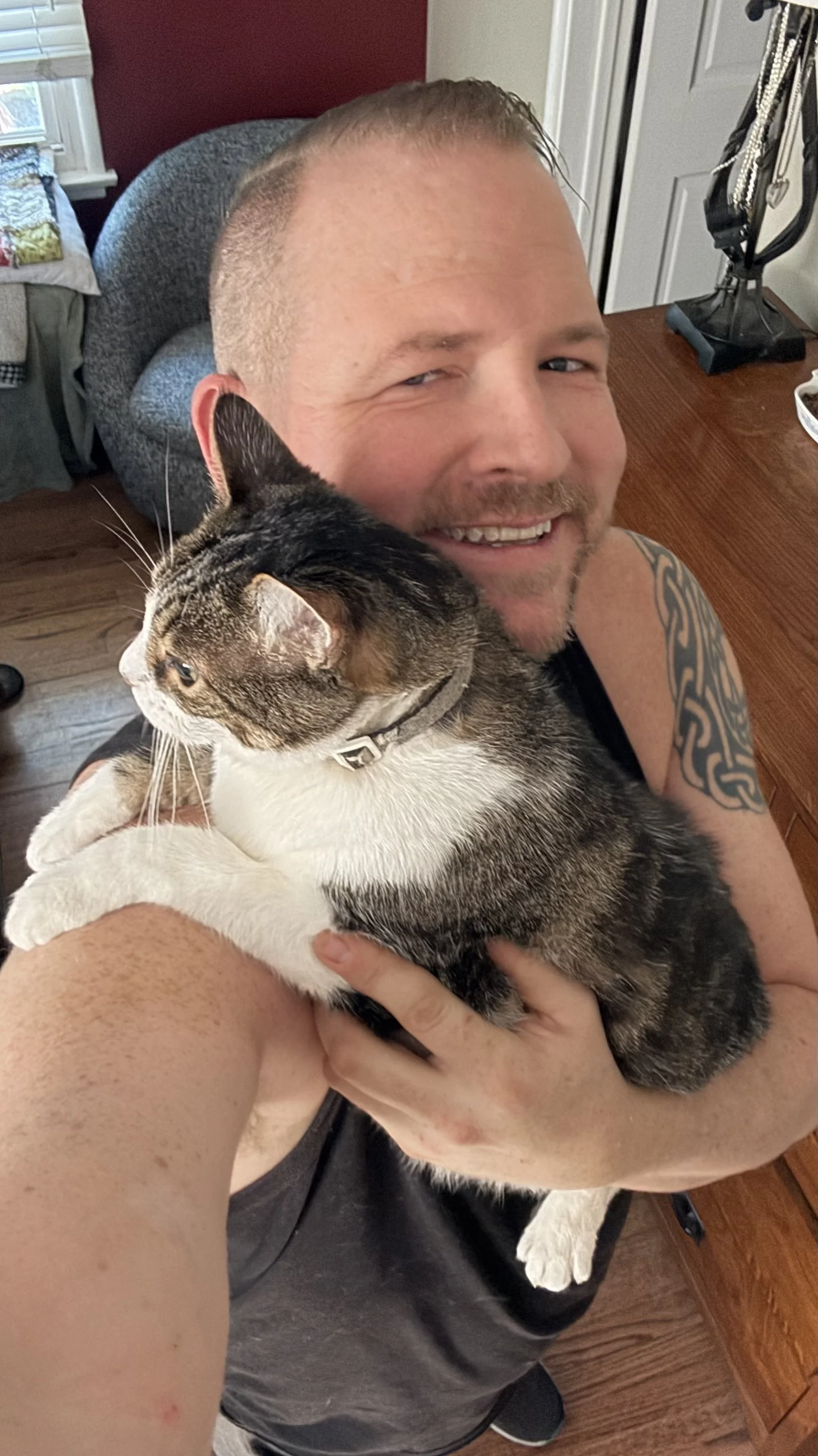 A man holding a cat while taking a selfie in a room with wooden furniture and a red wall.