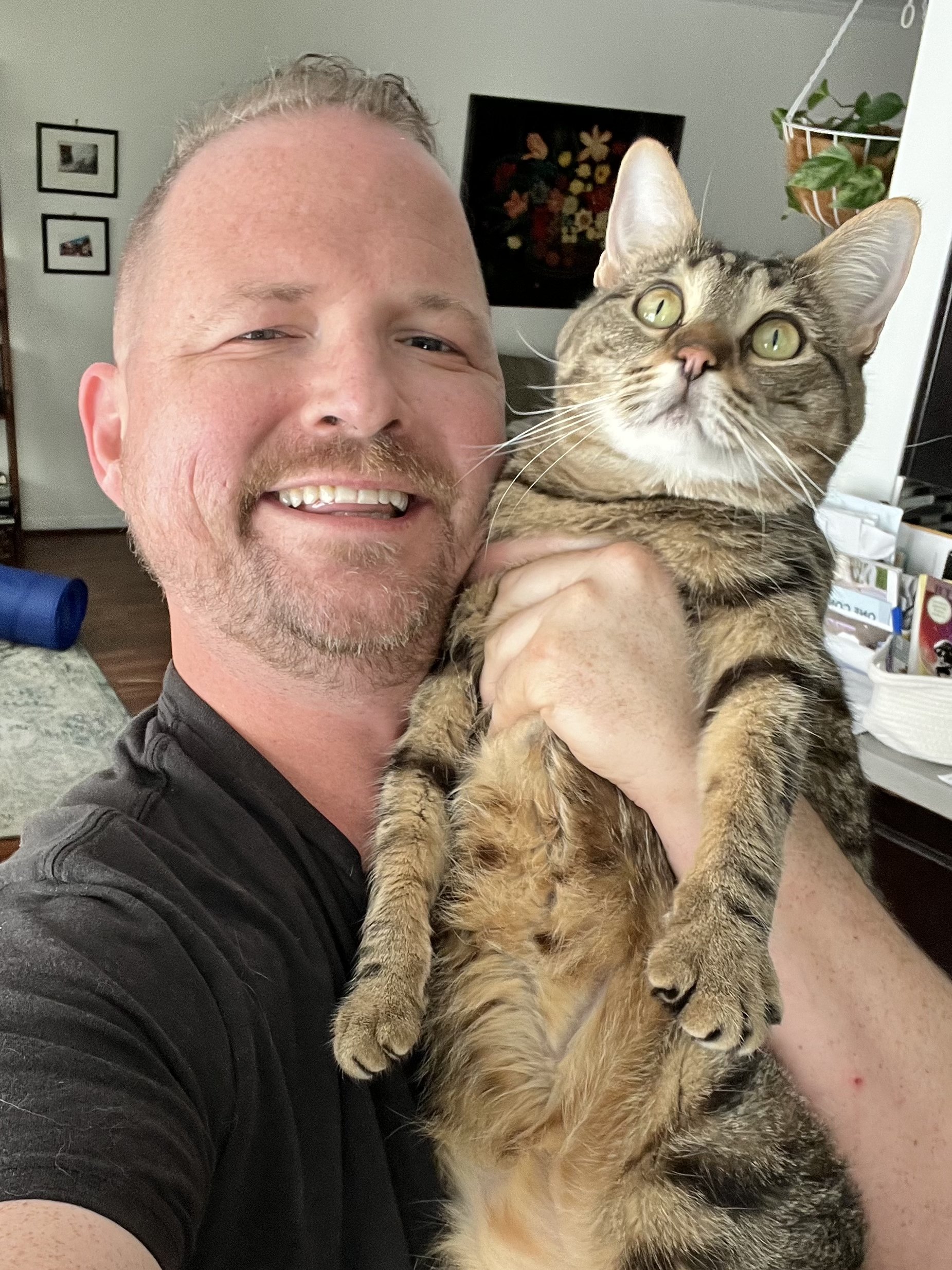 A smiling man holding a tabby cat with green eyes inside a home, with various pictures, plants, and household items in the background.