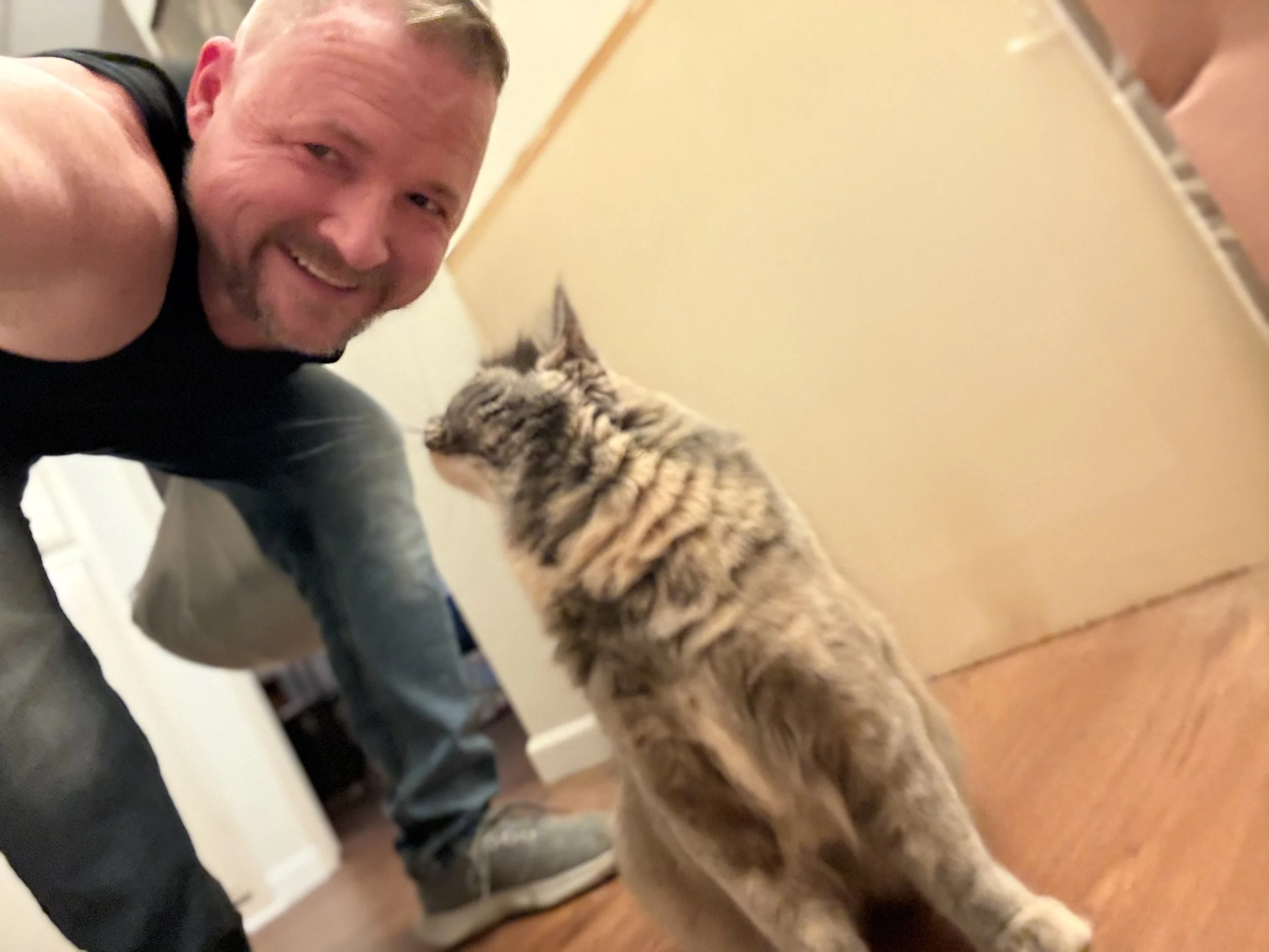 A man taking a selfie with a cat indoors. The man is smiling, wearing a black sleeveless shirt, and kneeling on the floor. The cat has a tabby fur pattern, and the background shows a hallway with beige walls and brown flooring.