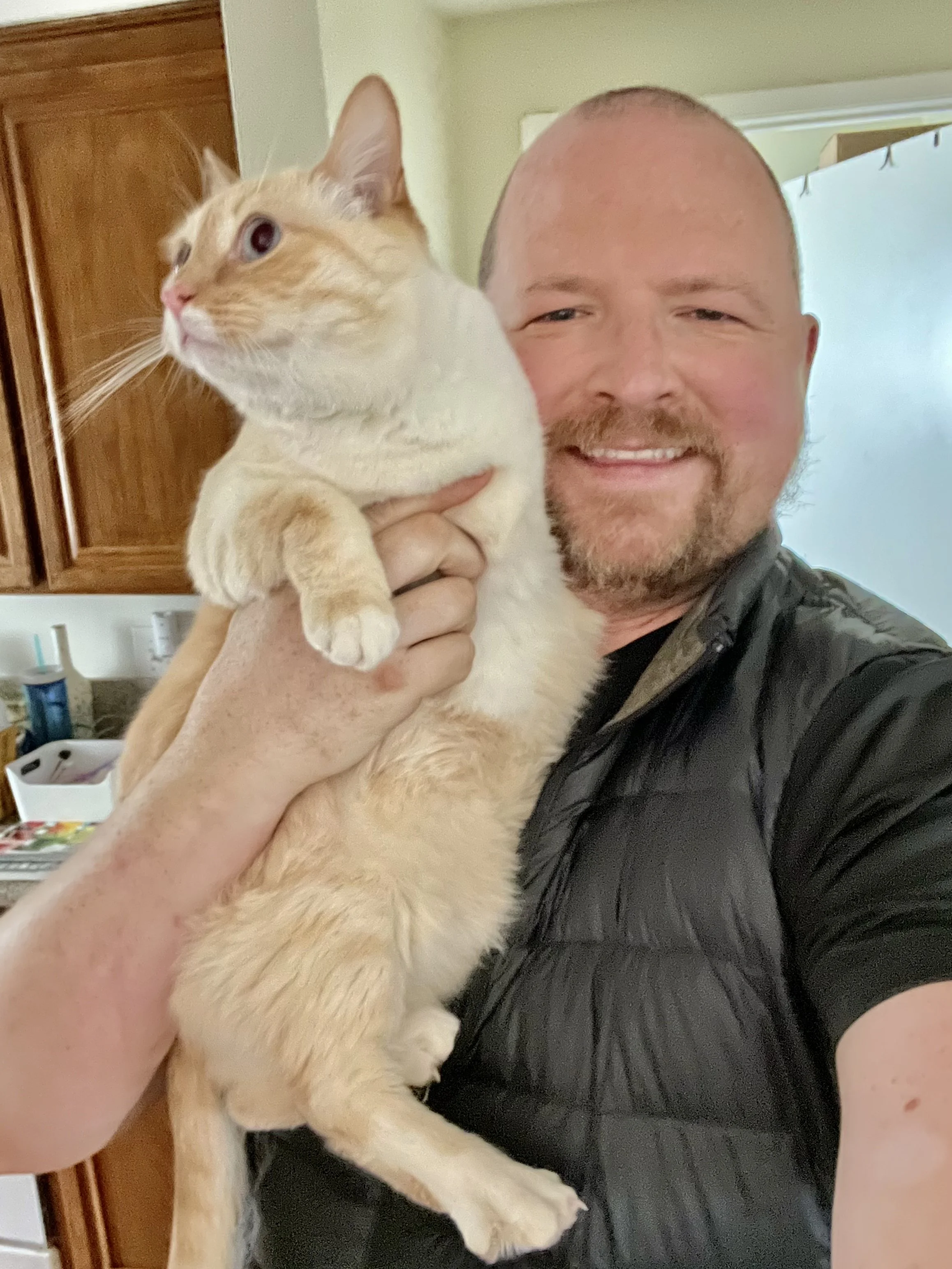 A smiling man holding an orange and white cat in a kitchen.