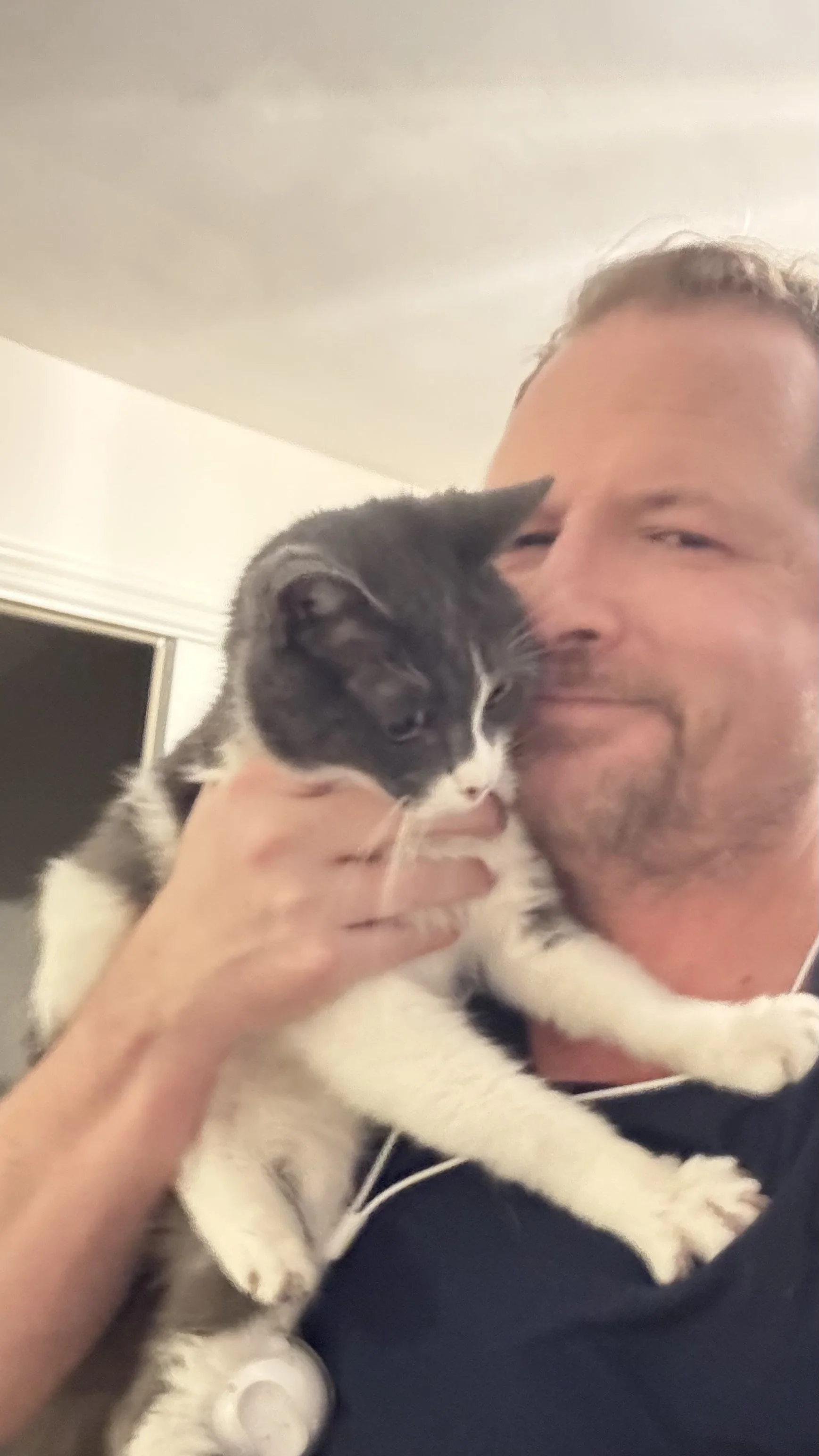 A man holding a black and white kitten close to his face, both appearing to enjoy a moment of affection indoors.