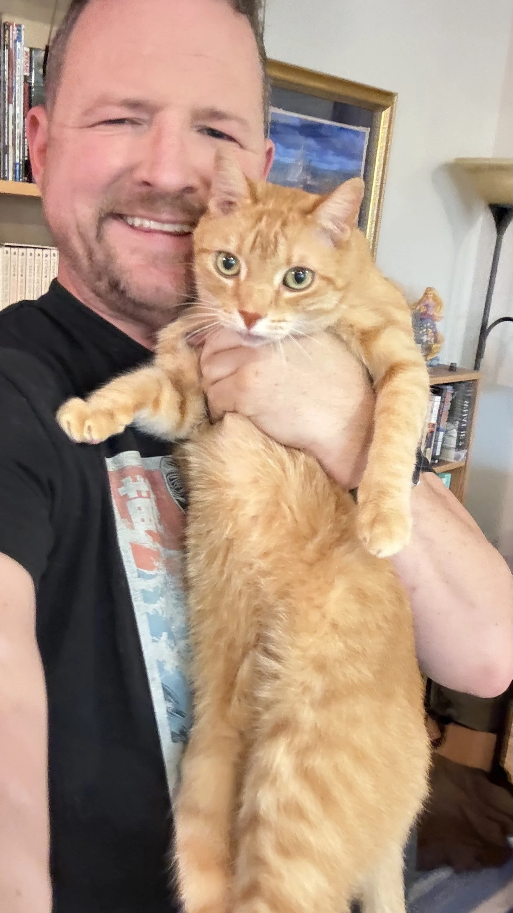 A man with a beard is smiling and holding an orange tabby cat, with a bookshelf and framed picture in the background.