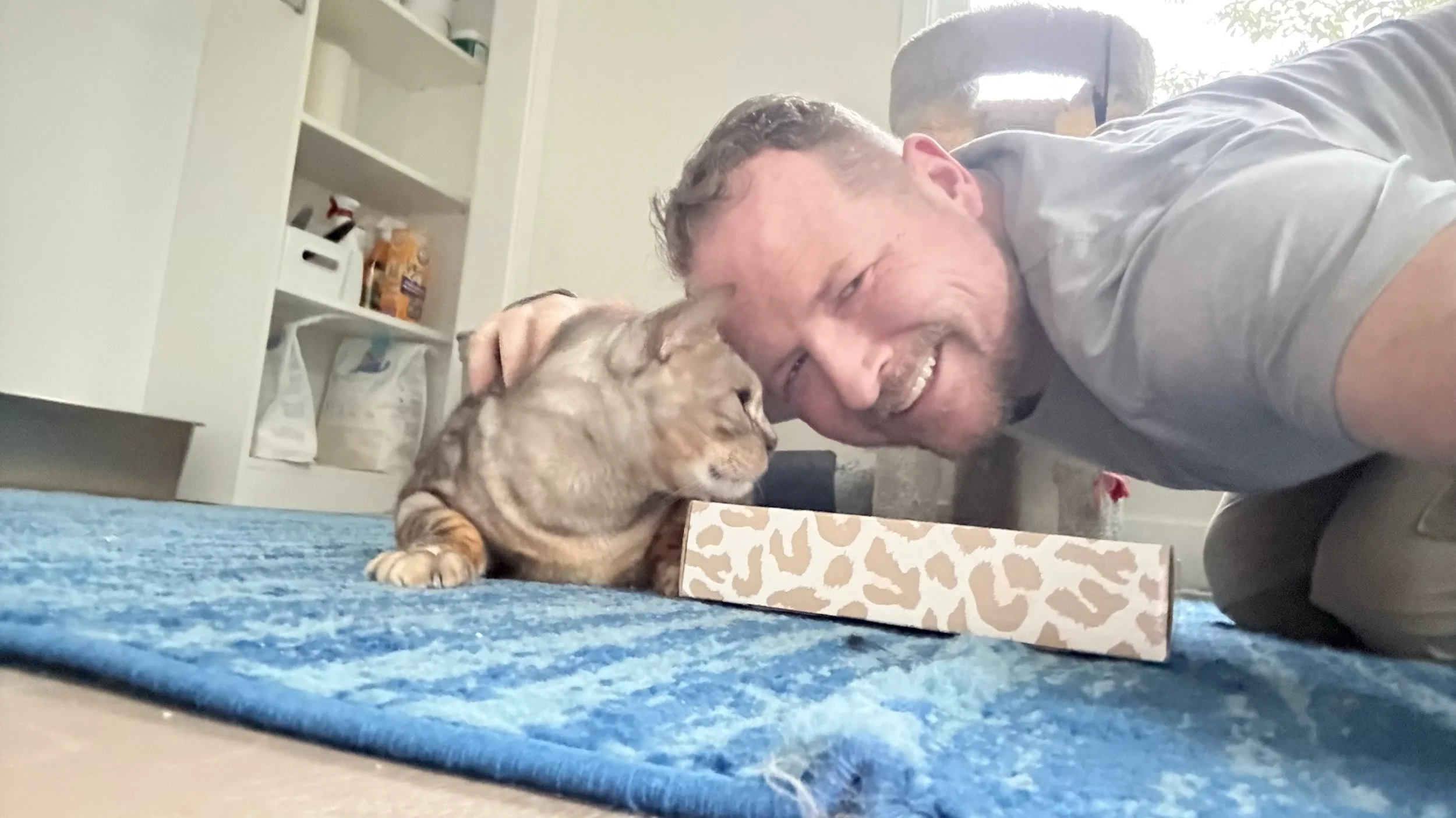 A man with a beard smiling, wearing a gray shirt, taking a selfie with a dog lying on a blue carpet and leaning on a leopard-print box. The man is bathed in natural light from a window behind him.