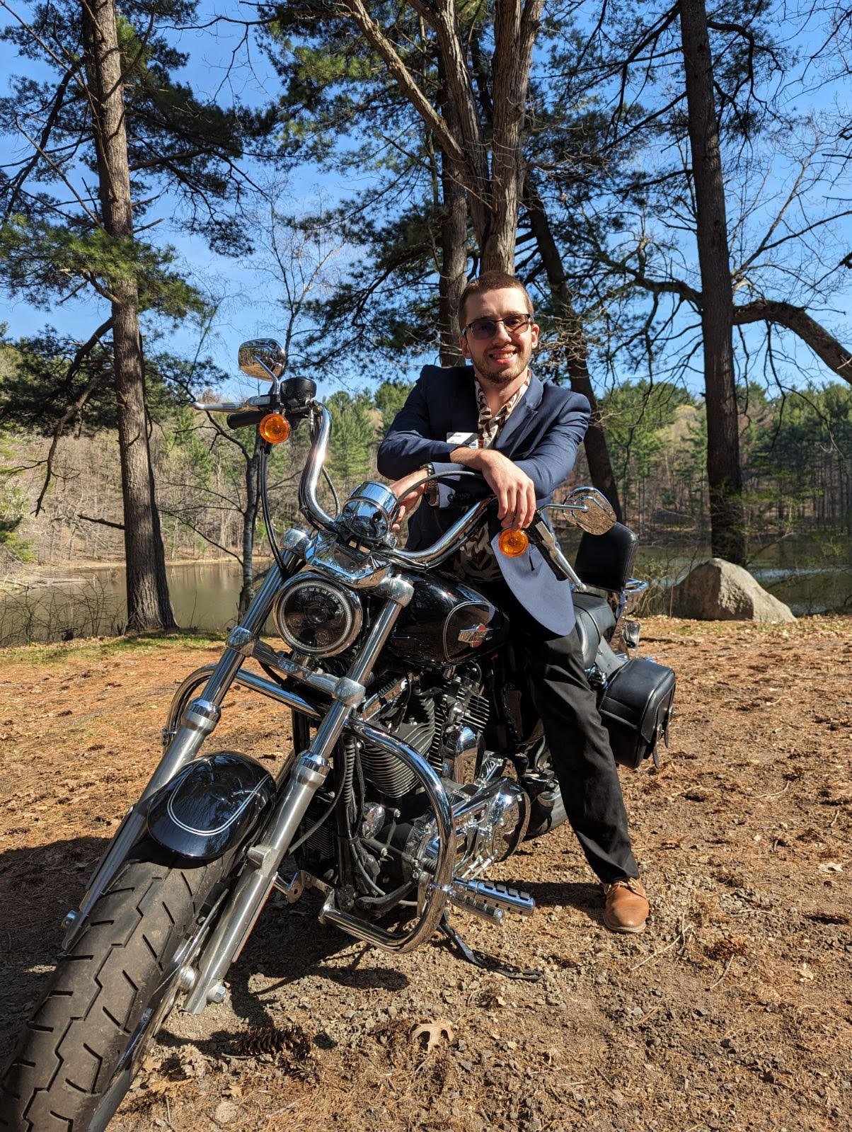 A man in a blue suit jacket and black pants sitting on a black motorcycle, smiling, outdoors in a wooded area near a lake with trees and rocks in the background.