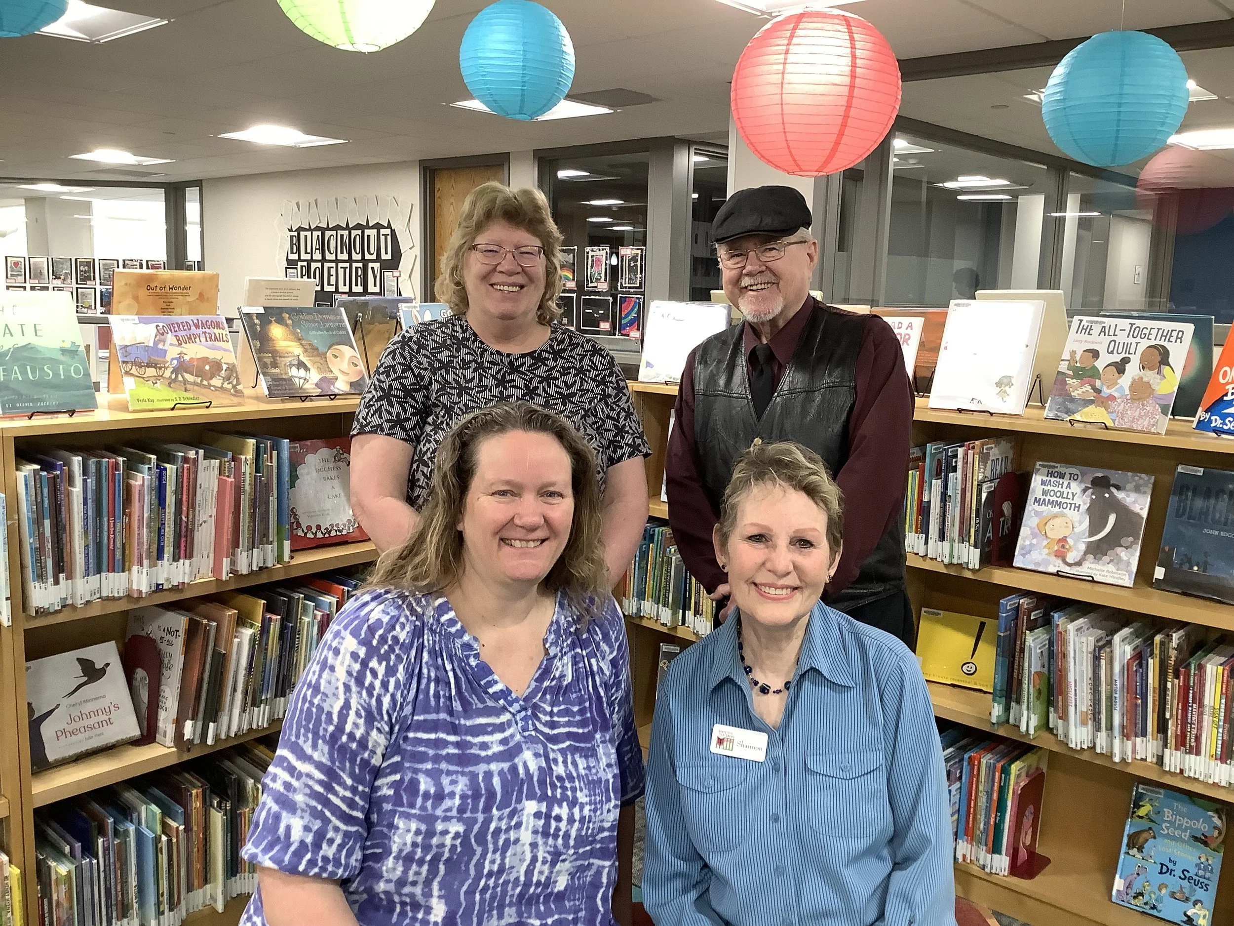 A group of four people standing in front of bookshelves in a library, with colorful paper lanterns hanging from the ceiling. The women are smiling and dressed in casual attire.