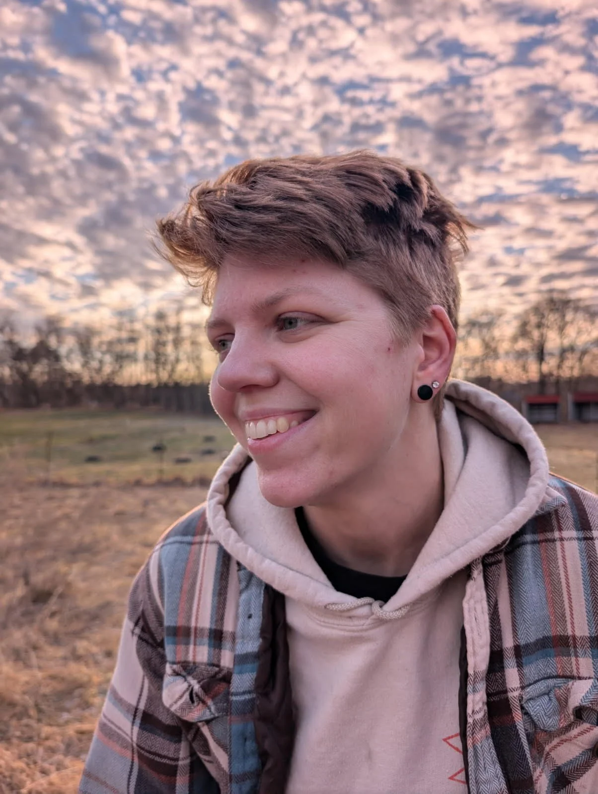 A smiling young person with short, styled hair, black earrings, and a plaid jacket, standing outdoors with a cloudy sky at sunset in the background.