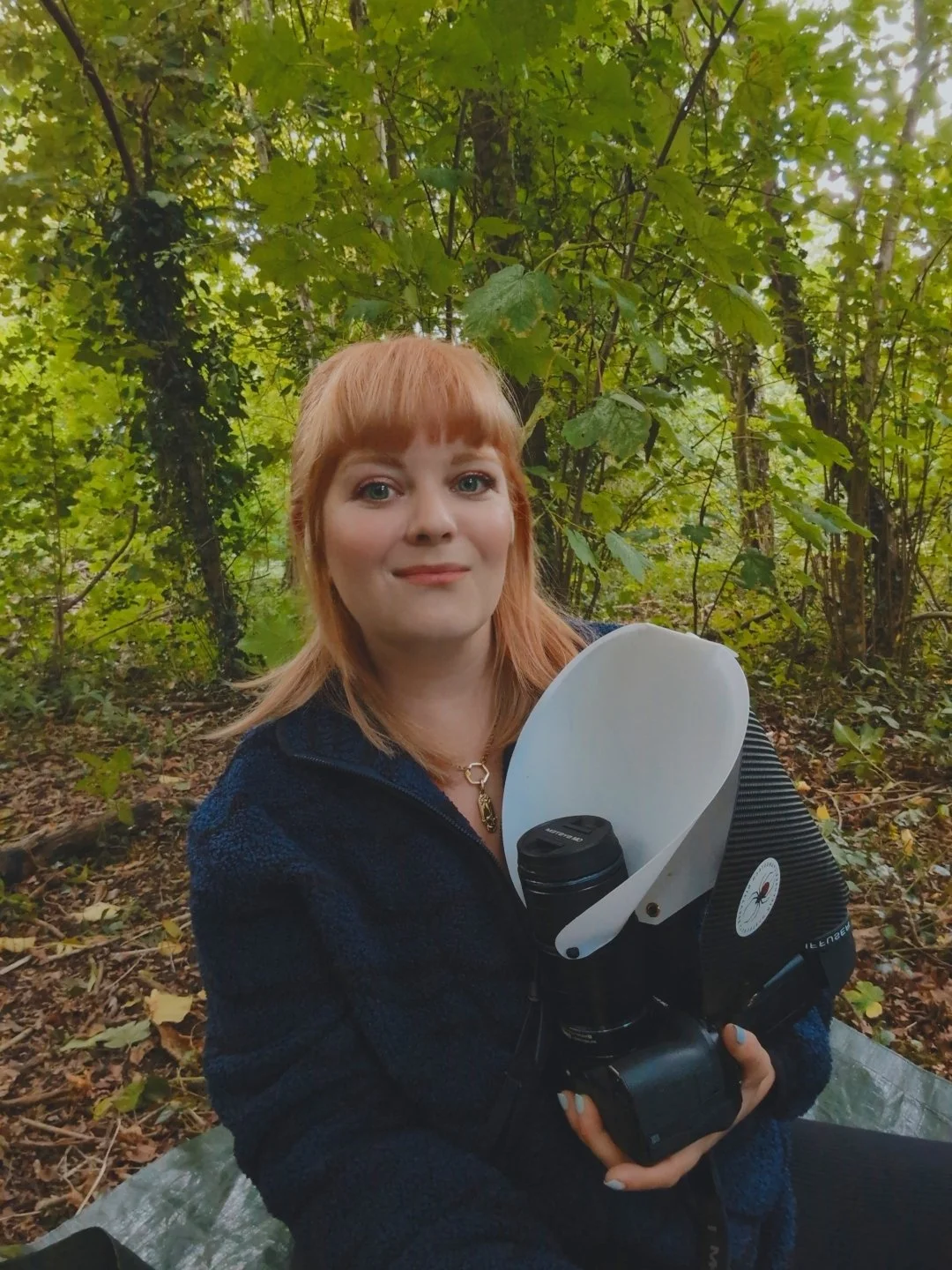 A woman with red hair holding a camera with flash diffuser in a forest with green leaves.