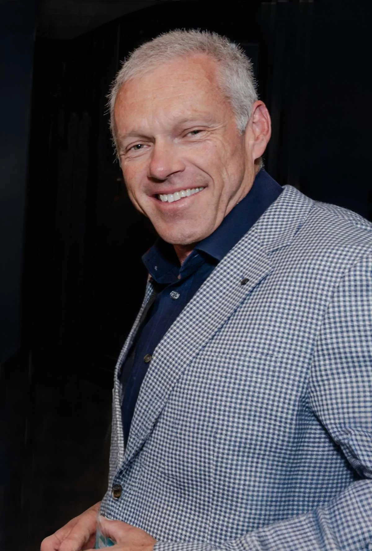 A smiling middle-aged man with short gray hair wearing a suit jacket and a navy blue shirt, standing against a dark background.