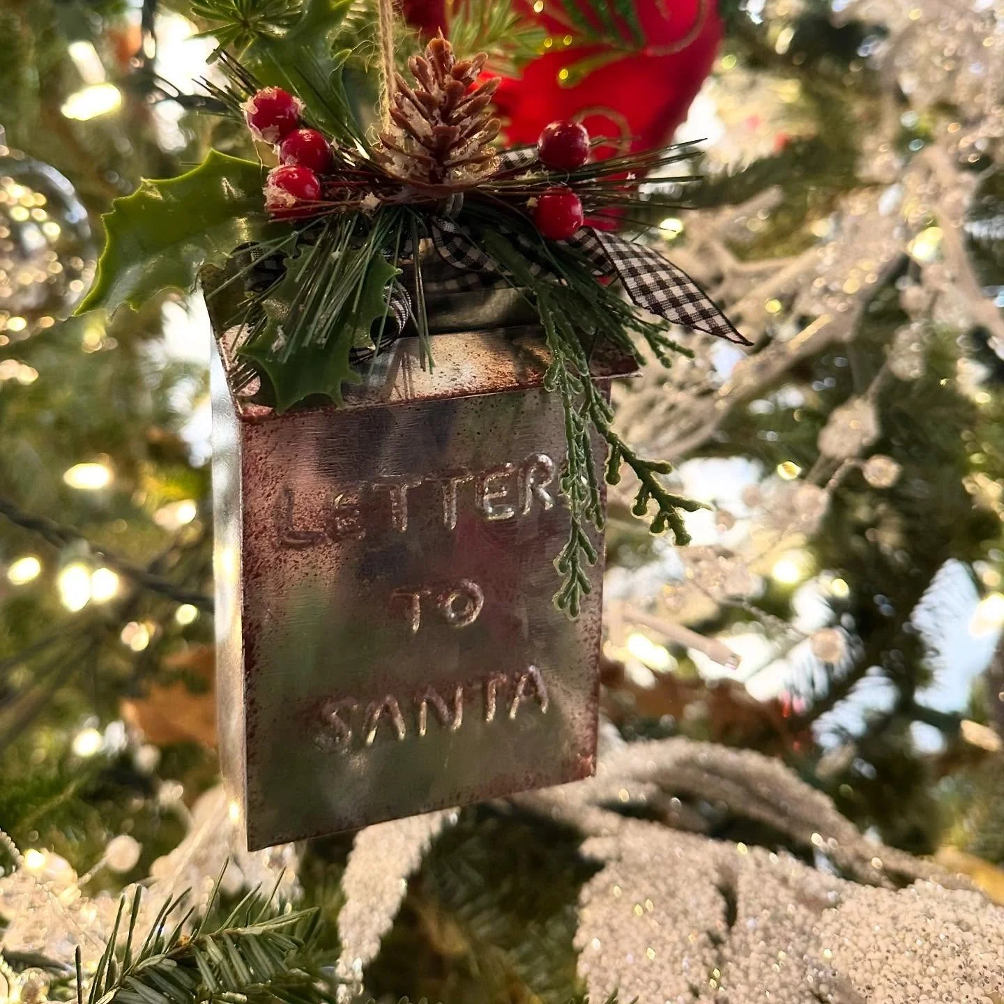 Close-up of a small festive ornament hanging from a Christmas tree branch, featuring holly leaves, red berries, pine cone, checkered ribbon, and a distressed metal tag with stamped text, with snow on the branches and blurred lights in the background.