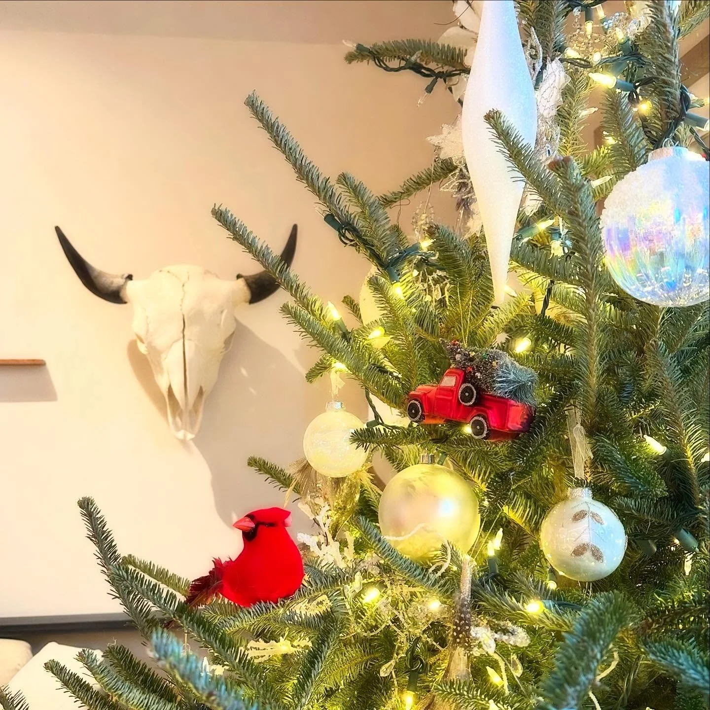 Decorated Christmas tree with ornaments and lights, a red toy truck, a red cardinal bird figurine, and a bull skull mounted on the wall in the background.