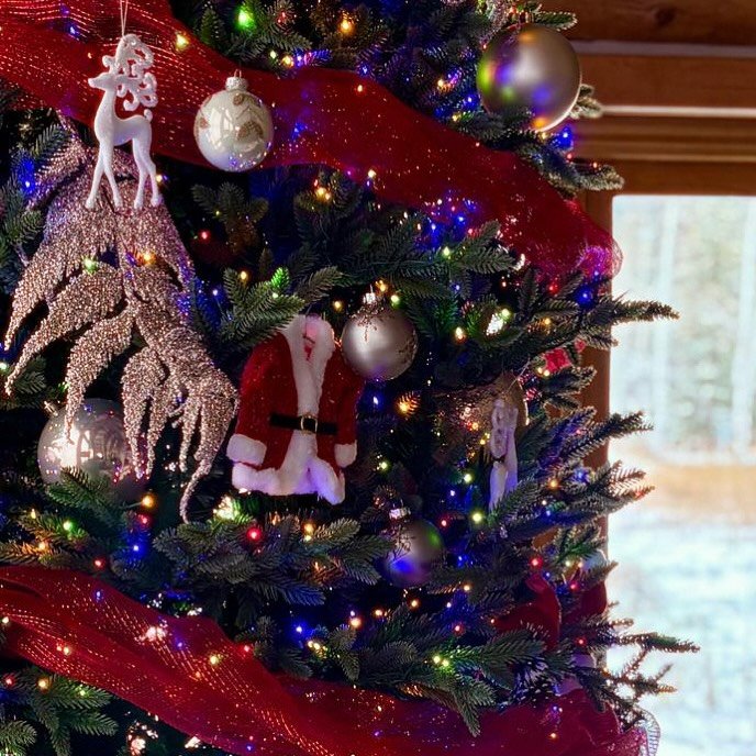 Close-up of a decorated Christmas tree with ornaments, ribbons, and lights in a cozy room.