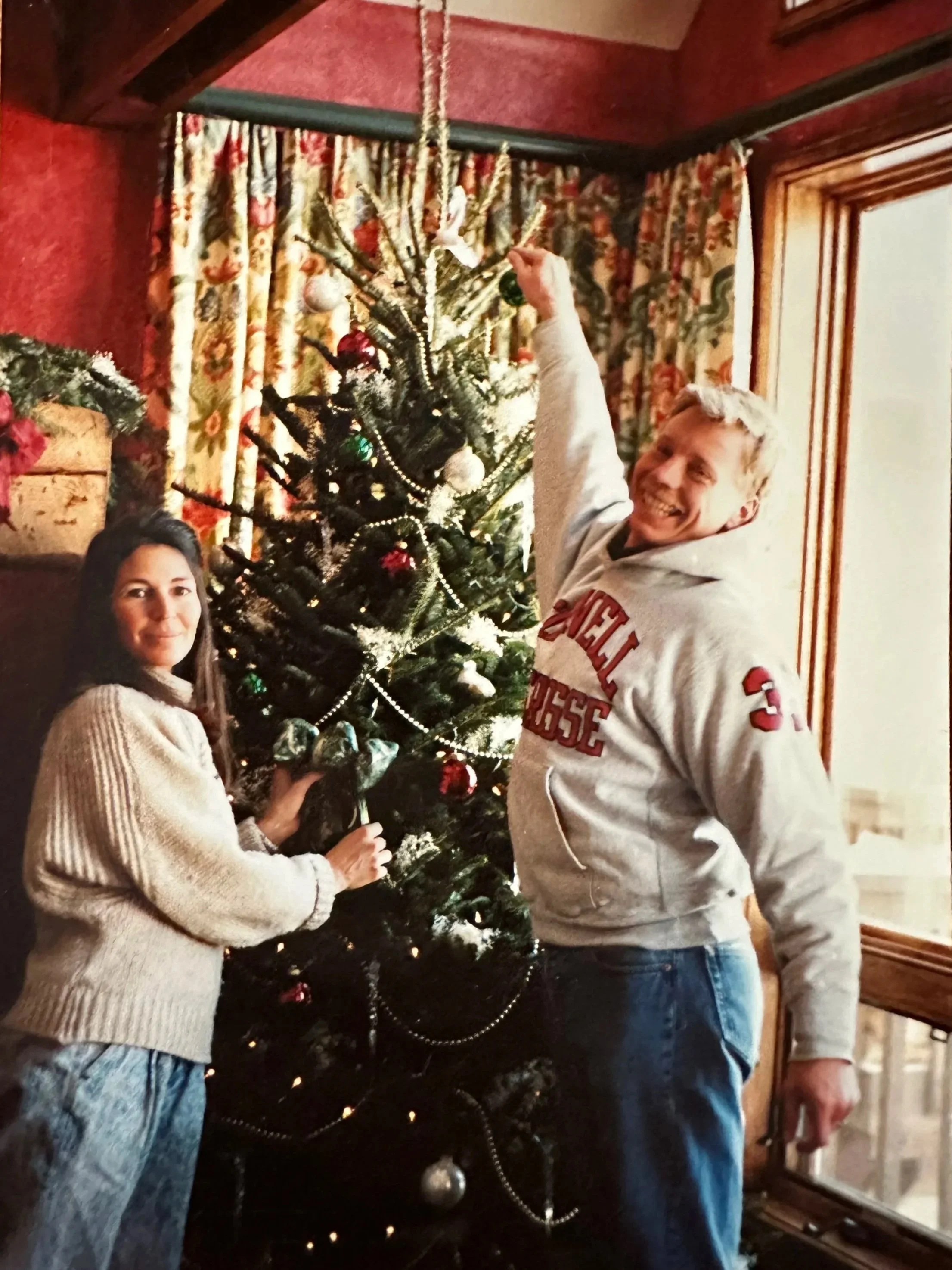 A woman and a man decorating a Christmas tree indoors. The woman is placing an ornament on the tree, and the man is smiling at the camera while holding an ornament. The Christmas tree is decorated with baubles, beads, and other ornaments. The room has red walls, floral curtains, and wooden trim.