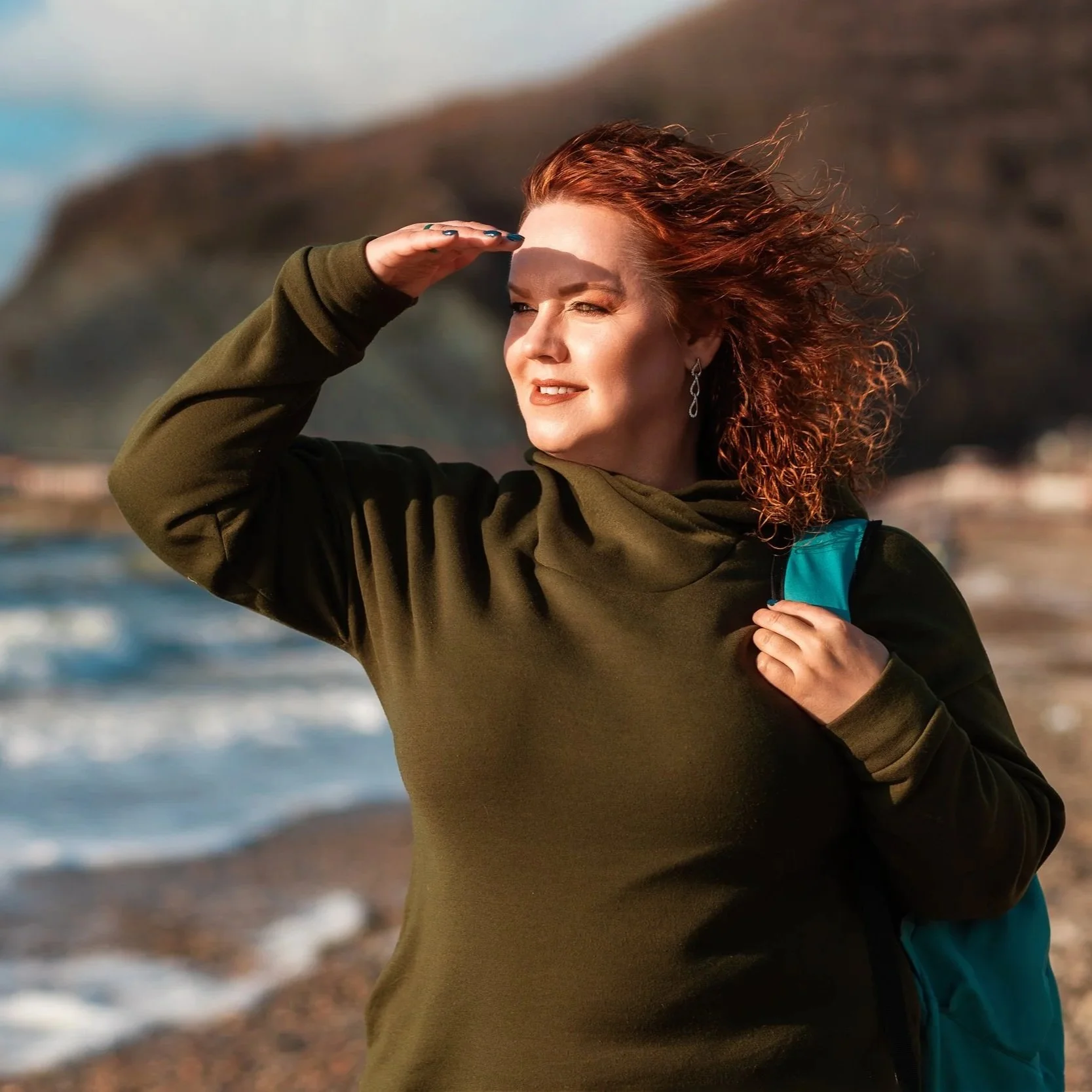 Woman with red hair and teal backpack on a beach looking to the distance