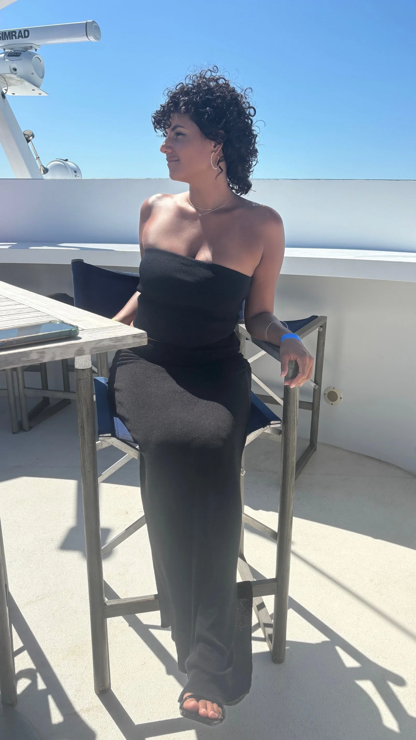 A woman with curly dark hair sitting on a barstool on a balcony or deck outdoor area, wearing a strapless black dress, jewelry, and sandals, with sunlight and a clear blue sky in the background.