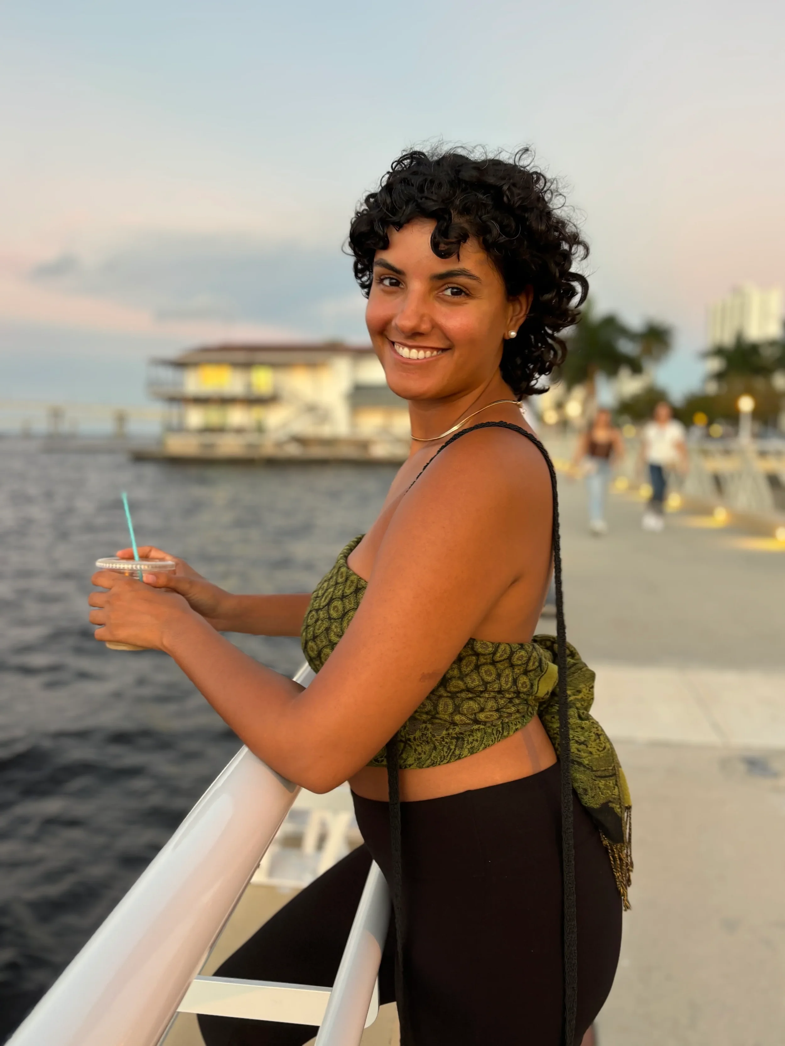 A woman with curly dark hair smiling on a boat, holding a drink with a straw, during sunset with a harbor and buildings in the background.