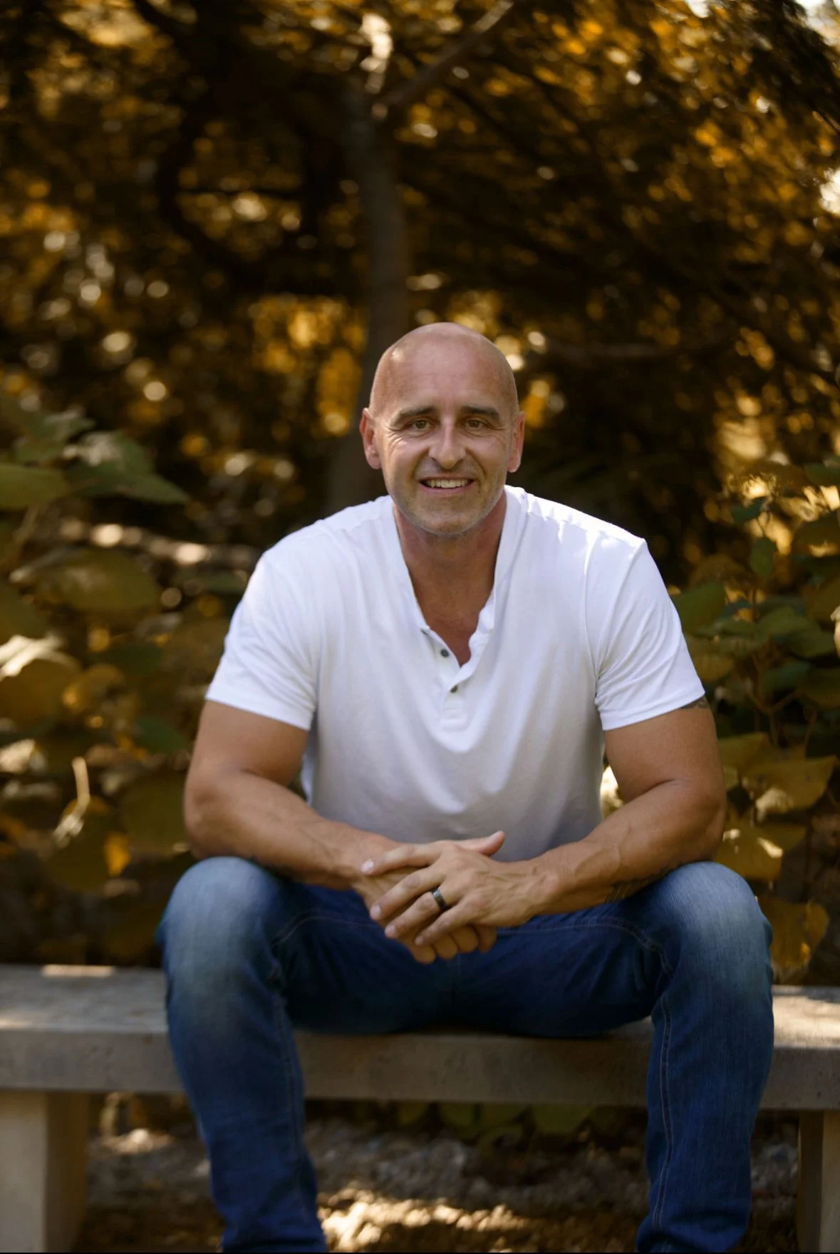 A bald man smiling and sitting on a bench outdoors in front of trees with golden leaves, wearing a white shirt and blue jeans.